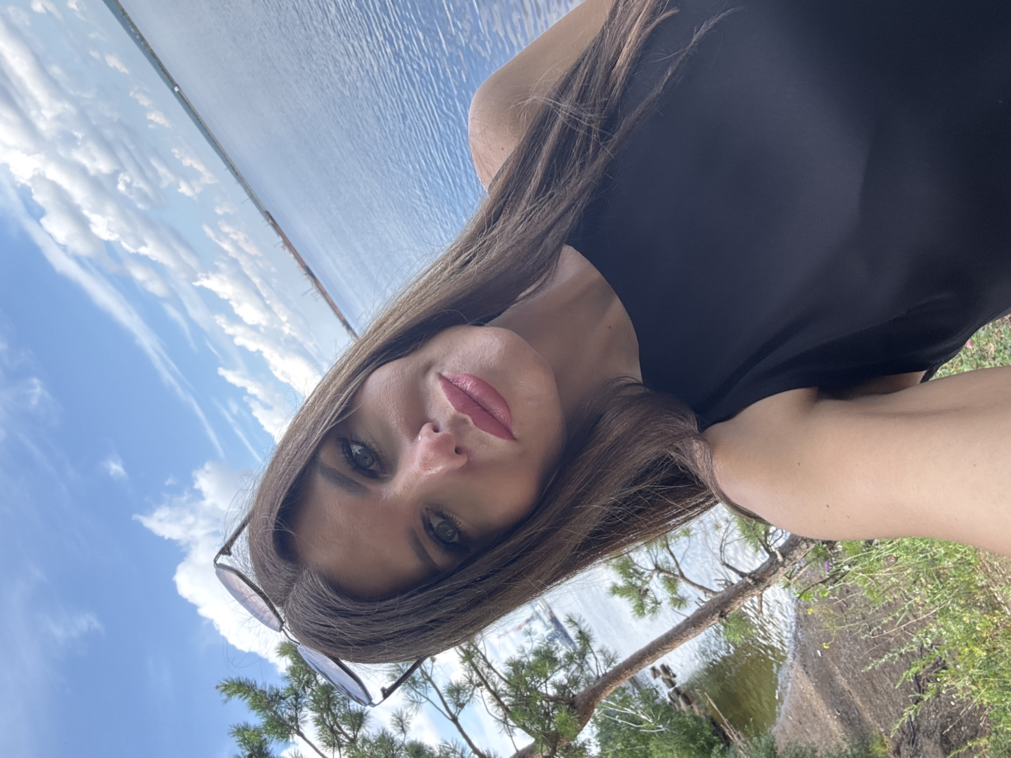A woman is taking a selfie by a body of water. The woman has long brown hair, wears a black top, and sunglasses are perched on her head. She is outdoors, with a body of water and a cloudy sky in the background. To the left, there are trees and foliage. The location is likely the Kama River embankment in Chaikovsky, Russia, as the river is wide and industrial structures are visible in the distance.