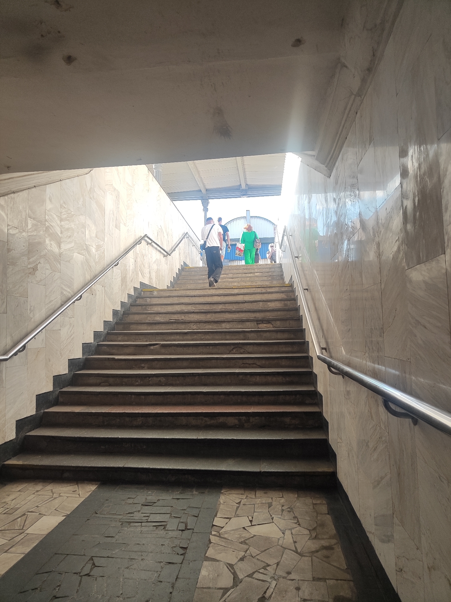 The image depicts a concrete and marble staircase in an underground passage. Several people are ascending the stairs, bathed in bright sunlight streaming from above. The scene is set in a public transit area, possibly a train station or subway, indicated by the overhead structure and the general architectural style.
The main subjects are the people walking up the stairs. One individual, wearing a white t-shirt and dark pants, is in the foreground, his back to the camera. Further up, a person in a vibrant green outfit is also moving upwards, carrying shopping bags. Other figures are visible in the distance, also heading towards the light.
The staircase is made of concrete treads with metal railings on both sides. The walls are clad in large marble tiles, reflecting the light. The floor at the bottom of the stairs is paved with a mosaic of irregular stone tiles. The overall impression is one of a functional, albeit somewhat aged, public space.
The bright sunlight filtering into the passage suggests it is daytime, likely late morning or early afternoon, given the angle of the light. The weather appears to be clear and sunny. There are no visible text elements in the image. The atmosphere is one of everyday movement and transition, with the light creating a sense of hope or arrival at the top.