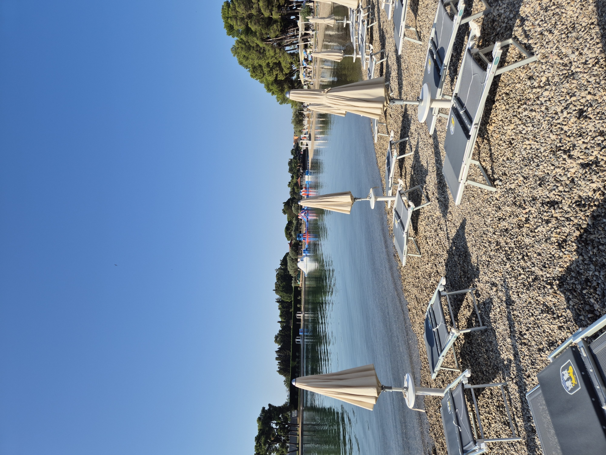This image depicts a tranquil scene at a beach resort in Umag, Croatia, characterized by a clear blue sky and calm, reflective water. The foreground is dominated by a pebbly beach adorned with numerous folded beach chairs and closed, light-colored umbrellas, suggesting an early morning or late afternoon setting when the beach is not yet crowded. These amenities are neatly arranged, indicating an organized resort environment.
The midground features a serene body of water, possibly a lagoon or a sheltered bay, where a variety of inflatable water park attractions are visible in the distance. These colorful structures hint at recreational activities available at the resort. The water's surface is smooth, reflecting the blue sky and the lush green trees that line the opposite shore.
The background is composed of a dense line of tall green trees, providing a natural backdrop to the resort. The trees are mature and varied in type, with some appearing to be pine trees, suggesting a Mediterranean or coastal climate. The overall impression is one of a well-maintained and inviting leisure destination, offering both relaxation and entertainment. There are no people visible in the frame, contributing to the sense of quietude and anticipation.
