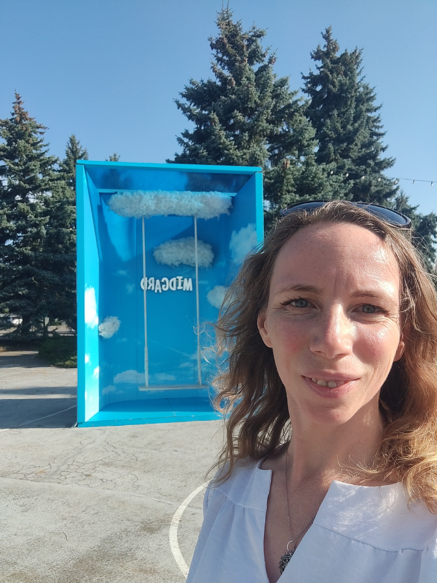 A woman with shoulder-length auburn hair and blue eyes is taking a selfie outdoors. She is smiling, and wearing a white v-neck shirt and a silver necklace. Behind her is a bright blue rectangular installation, resembling a box, with fluffy white clouds and a swing inside. Text on the back wall of the installation reads "WIDCMAD" in white letters, which is "MADMCIDW" spelled backward. The installation appears to be set up on a paved surface with white line markings. In the background, several tall evergreen trees stand against a clear blue sky. The setting suggests an outdoor public space or park. There is no visible indication of this being in Kyiv, Ukraine.