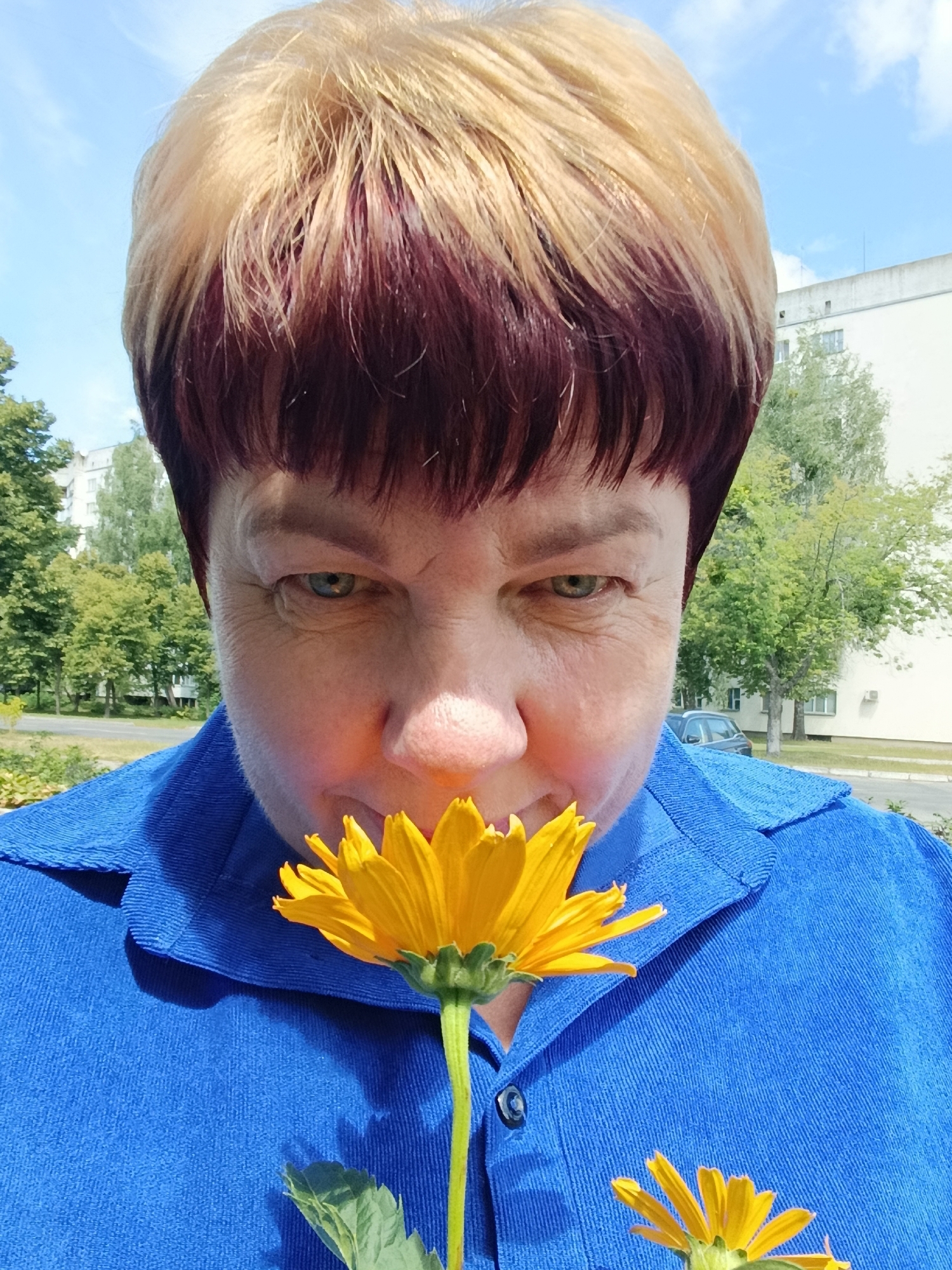 The image captures a close-up, selfie-style portrait of a woman with short, two-toned hair (blonde on top, burgundy underneath) against an outdoor, urban backdrop. She is wearing a blue corduroy collared shirt. Her eyes are green, and she is leaning in to smell a bright yellow flower held close to her face. Her nose is visible, and her mouth is slightly obscured by the flower. The background shows a paved area, green trees, a light-colored building, and a blue sky with scattered clouds, consistent with a location in Svietlahorsk, Belarus. There is another smaller yellow flower visible at the bottom right of the frame.