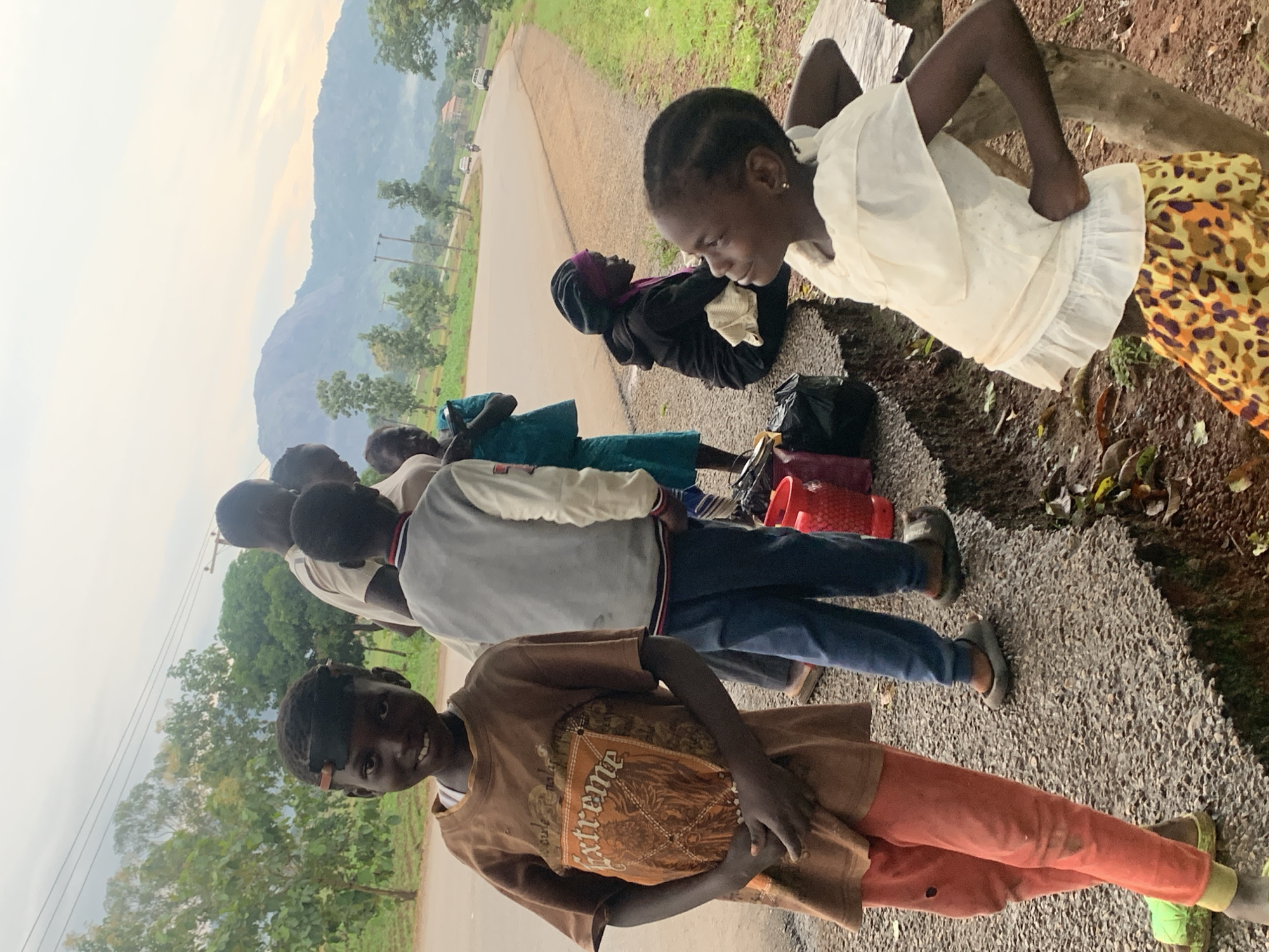 This image captures a group of people, primarily children, gathered outdoors on a roadside in Agban, Nigeria. The foreground features a young boy with dark skin and hair, wearing a brown t-shirt with an "Extreme" graphic and orange pants. He stands with his hands clasped in front of him, looking towards the camera with a slight smile. To his right, another boy in a grey jacket and blue pants stands partially obscured.
In the mid-ground, a young girl in a teal dress is seen holding something, and further back, a woman with a dark headscarf sits on a concrete ledge. Beside her, a girl in a white top and a patterned skirt is looking upwards and to the right, her hands on her hips. A collection of red buckets and black bags are visible on the ledge near the sitting figures.
The environment suggests a rural or semi-rural setting. A paved road curves in the background, with a few vehicles, including cars and a motorcycle, visible on it. Rolling hills with lush greenery and scattered trees dominate the landscape, with a large mountain range visible in the distance under a cloudy sky. The ground in the foreground consists of gravel and a section of broken concrete embankment. The overall lighting indicates it might be early morning or late afternoon, given the soft light and overcast conditions.