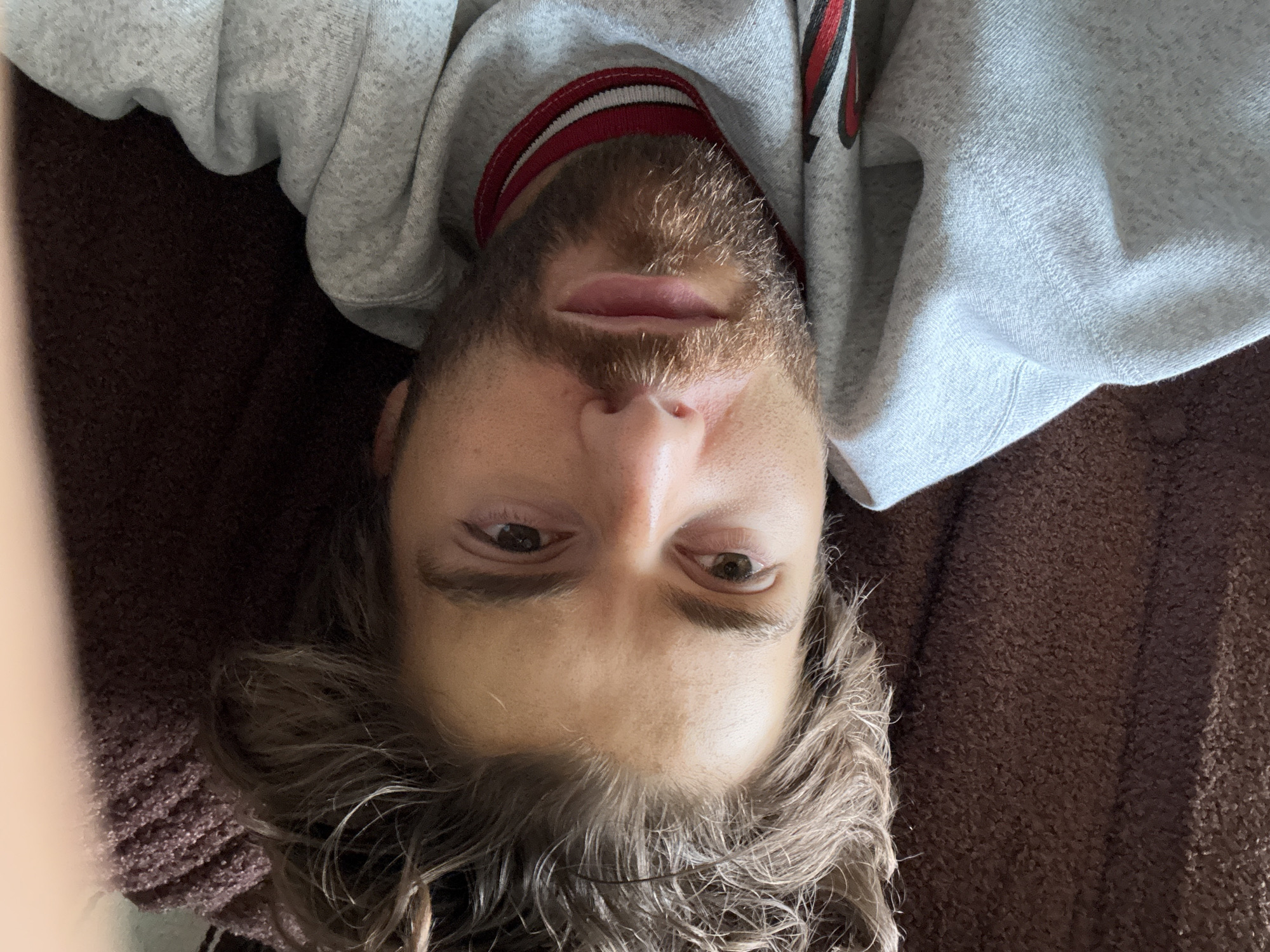 A close-up, upside-down selfie shows a person with light-brown eyes, light-brown hair, and a beard. They are wearing a grey hoodie with a red and white collar. The person is lying on a brown, textured surface, possibly a sofa or bed. The location is unknown and cannot be deduced from the image. The city of Montreal, Canada cannot be deduced from the image.