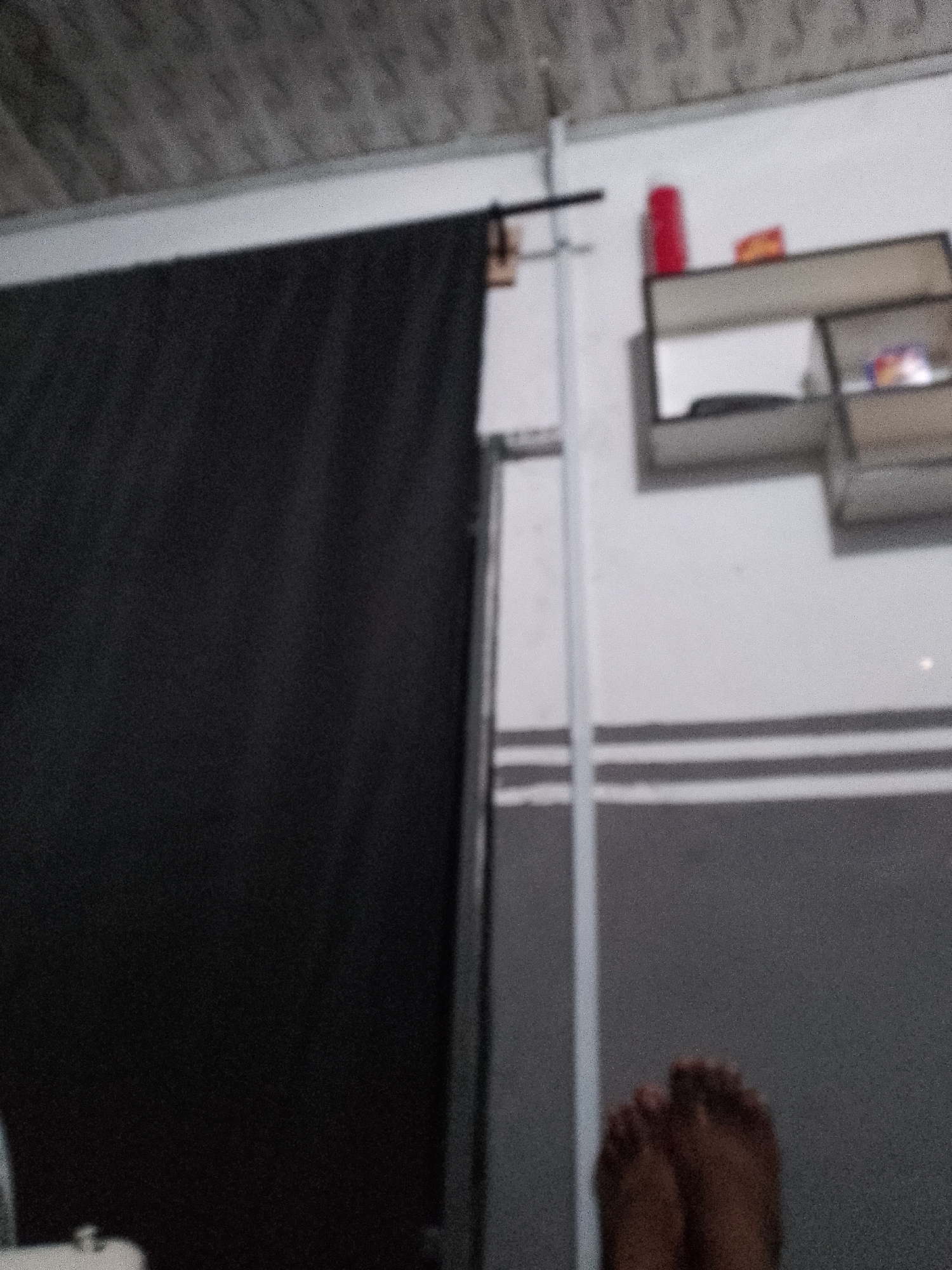 The image shows a dimly lit interior scene. On the left is a large black curtain or fabric. To the right, a white wall has a grey horizontal stripe pattern. Above the stripe is a grey shelf unit with some blurred items on it, including a red cylindrical object and what appear to be packaged goods. In the foreground, the lower legs and feet of a person are visible, resting on a grey surface. The overall impression is of a casual, possibly domestic, setting in Jauro Yinu, Nigeria.