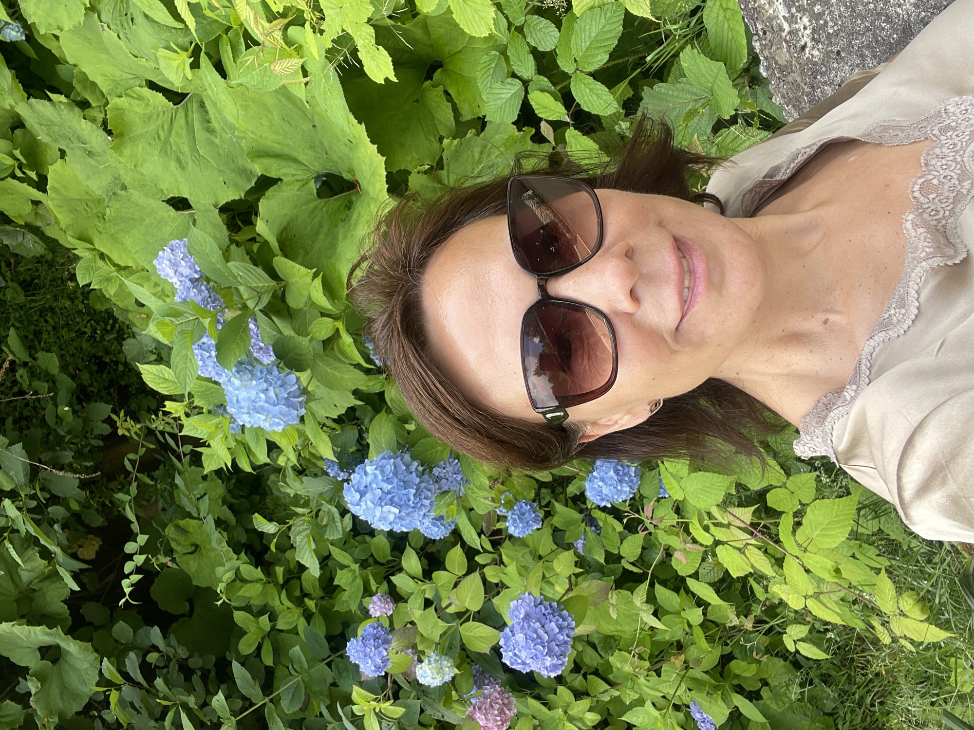 In Tqwarchal, Abkhazia, a woman with brown hair and sunglasses takes a selfie in front of a lush green backdrop filled with blue and pink hydrangeas. She is wearing a light-colored top with lace trim. The woman is smiling and looking at the camera. The environment appears to be a garden or park, with dense foliage and flowers creating a vibrant, natural setting. The lighting suggests it is daytime, possibly overcast given the soft light on her face.