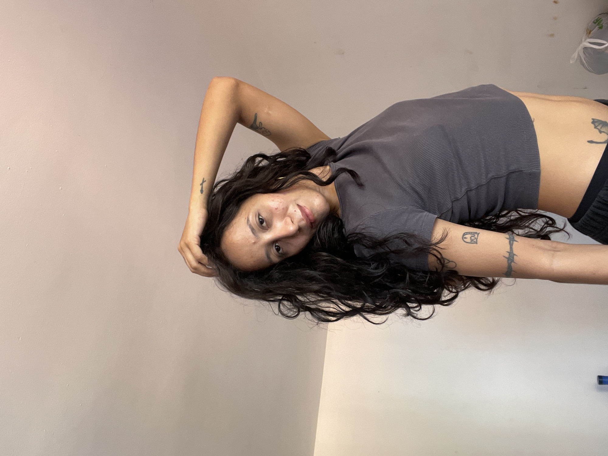 In the city of Porlamar, Venezuela, an individual with long, dark, curly hair and fair skin is captured from the waist up, leaning towards the camera. They are wearing a grey, ribbed, crop top that exposes their midriff, revealing a tattoo on their lower abdomen. The individual's left arm is bent at the elbow, with their hand resting on the back of their head, and their right arm is raised, with their hand touching their head. Tattoos are visible on both arms. The background consists of a plain, light-colored wall and ceiling, suggesting an indoor setting. The lighting is soft, casting subtle shadows.