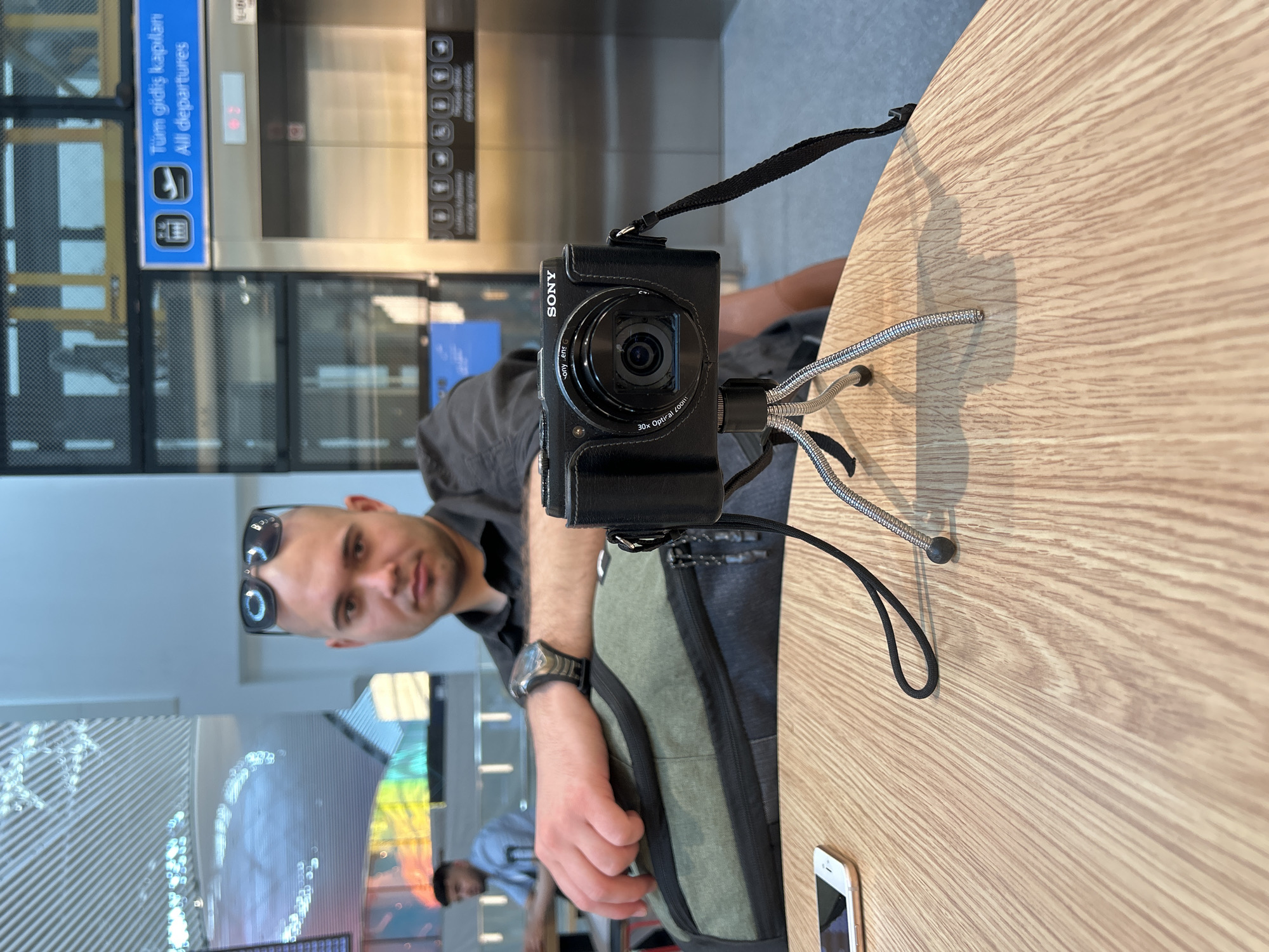 This image depicts an indoor scene, likely at a modern airport or transit hub in Arnavutköy, Turkey. A man, appearing to be in his late 20s or 30s, with short dark hair and sunglasses resting on his head, is seated at a light-colored wooden table. He is wearing a dark shirt and a watch on his left wrist, with an olive-green backpack resting beside him. He looks directly at the camera with a neutral expression.
In the foreground on the table, a black Sony compact camera is mounted on a small, flexible silver-legged tripod, its lens pointed towards the viewer. A white smartphone is also visible on the table to the right.
The background features contemporary architecture with large glass panels and metal structures, possibly elevators or entryways. A prominent blue sign in the upper left corner displays "Tüm gidiş kapıları" (Turkish for "All departures") and "All departures" in English, along with icons of an airplane and luggage, confirming an airport environment. Another person is faintly visible in the deeper background. The lighting suggests it is daytime.
Visible text includes "Tüm gidiş kapıları" and "All departures" on the blue sign. On the camera, "SONY," "30x Optical Zoom," and partially "Sony Lens G" are legible.
