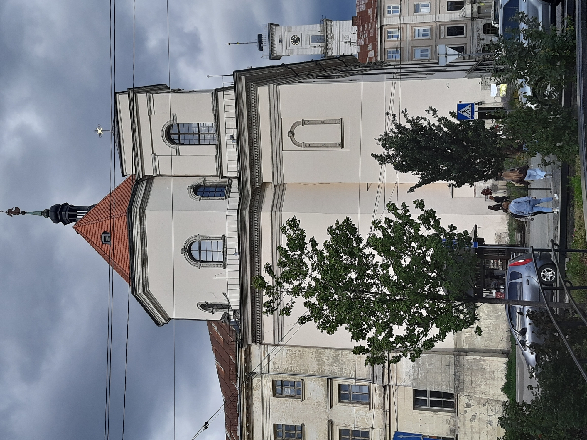 This image depicts a view of the Church of the Transfiguration in Lviv, Ukraine. The church, with its distinctive white facade and red tiled roof, is a prominent feature of the scene. A tall clock tower, likely part of another significant building, stands in the background to the right. Overhead, a network of power lines crisscrosses the sky, which is filled with dramatic, dark clouds, suggesting an overcast or stormy day.
In the foreground, a silver car is parked on the street. Several people are visible walking on a sidewalk in front of the church. Two young women, one in a denim jacket and the other in a brown jacket, are walking away from the camera. Further down the sidewalk, a group of people are walking towards the camera, partially obscured by trees. Lush green trees and bushes line the sidewalk and partially obscure the lower portion of the buildings. The architecture of the surrounding buildings is typical of Lviv, with multiple stories and varied window designs. The overall impression is of a bustling urban environment with historical architectural elements under a dynamic sky.