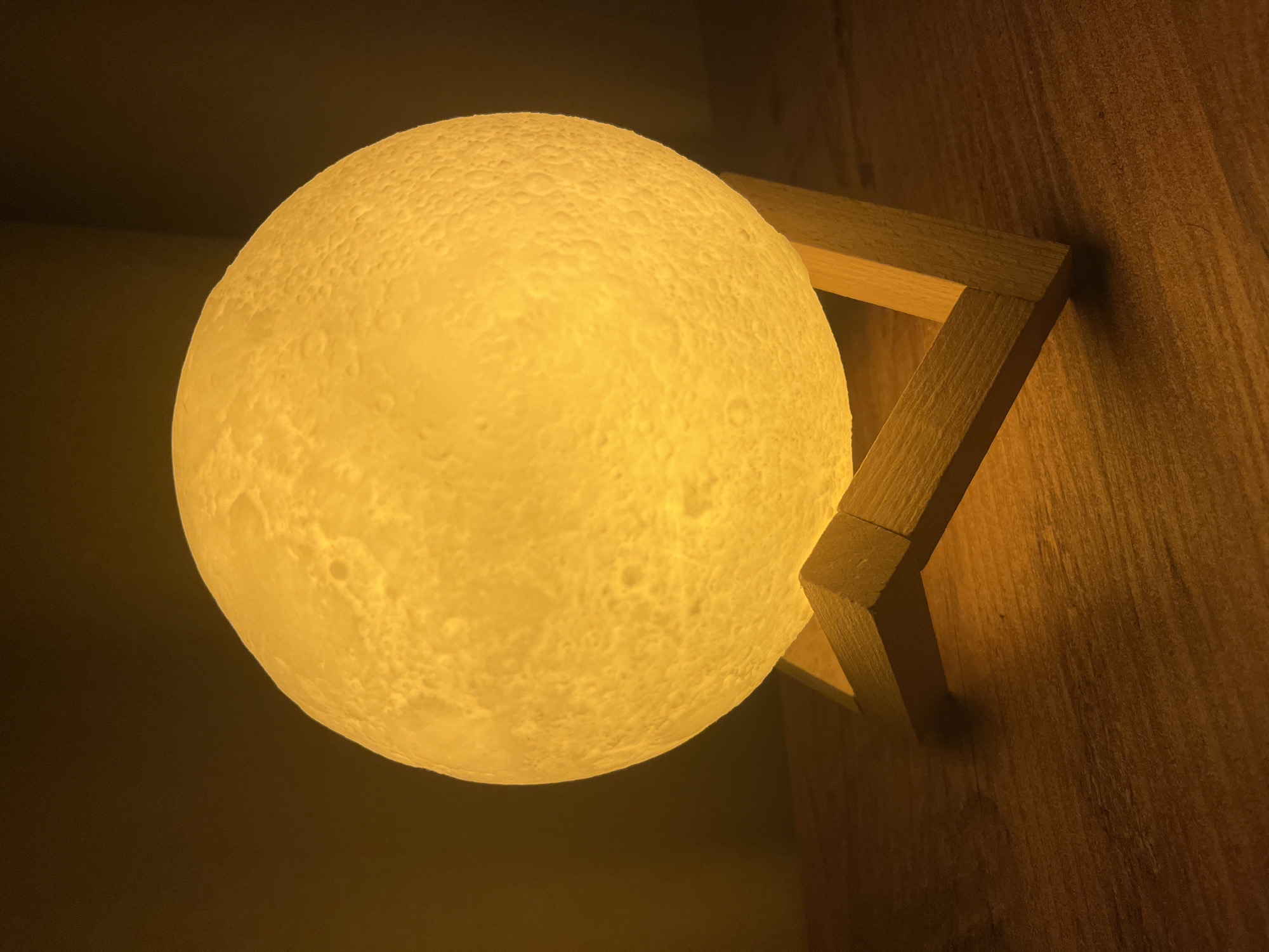 A close-up shot depicts a glowing, spherical lamp resembling the moon. It is mounted on a wooden bracket that is affixed to a wooden wall. The environment is dimly lit, emphasizing the warm glow of the lamp. The location is unknown.