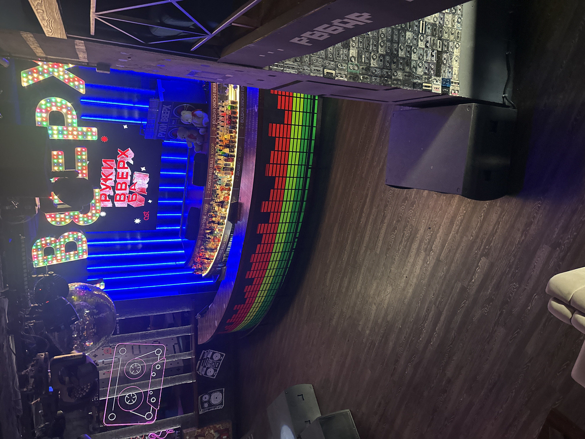 This image captures an interior view of the "Ruki Vverkh Bar" (Руки Вверх Бар) nightclub located in Moscow, Russia. The bar features a retro-themed environment with elements like a disco ball, neon signage spelling out the bar's name in both English and Russian, and a display of audio cassettes. The layout includes a well-stocked bar, colorfully lit, with a digital display screen on the front facade. The floor has a wood-grain pattern and there's a large audio speaker on the right side. A glimpse of seating is visible on the right edge, and various decorations and lighting fixtures are scattered throughout.