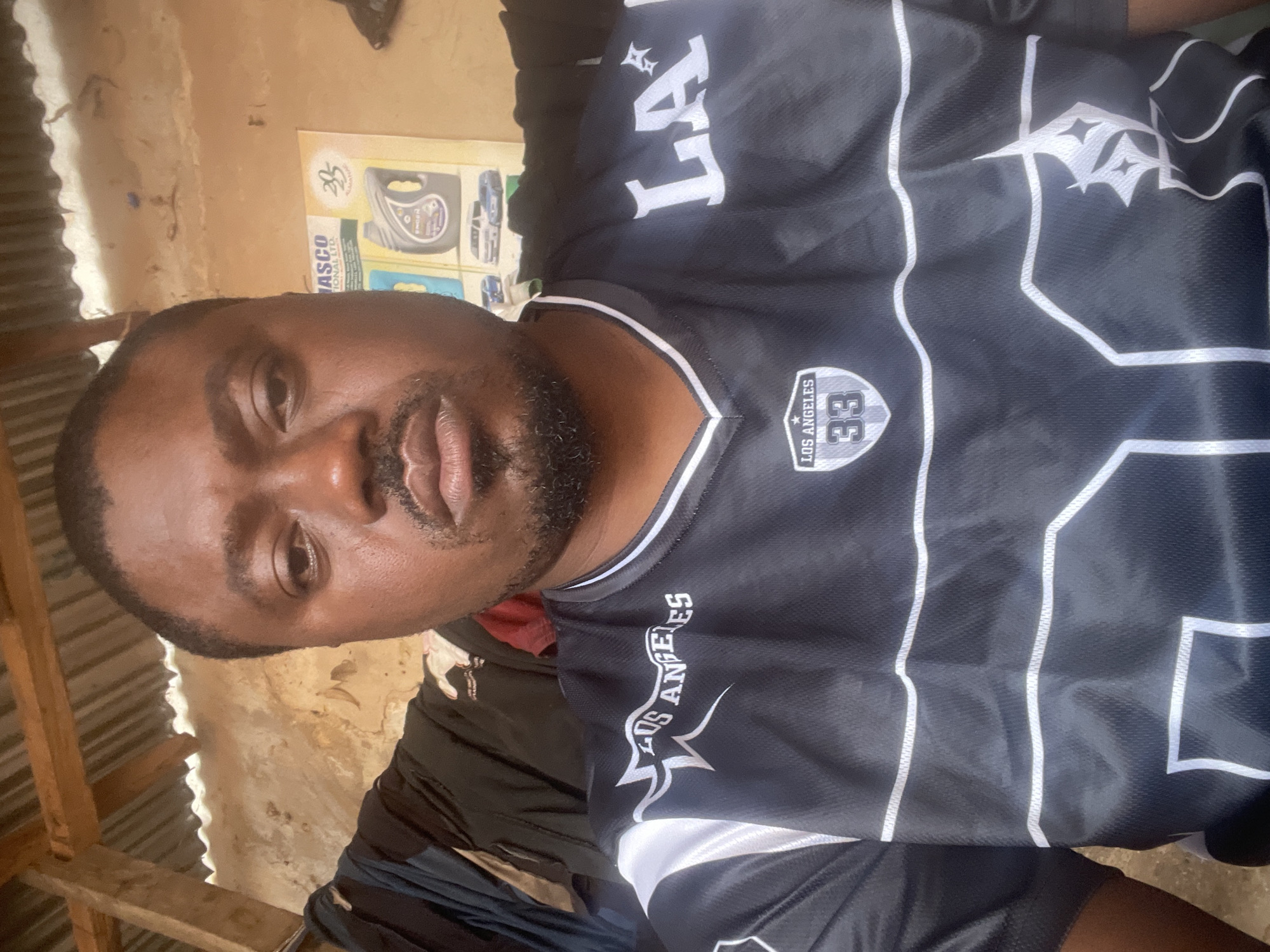 A middle-aged Black man is seen in a selfie, wearing a black and white "Los Angeles 33" jersey. The background shows a corrugated iron sheet wall, and a poster of motor oil. The location is claimed to be in Jos, Nigeria. The man has a beard and appears to be indoors, given the roofing and wall structure.