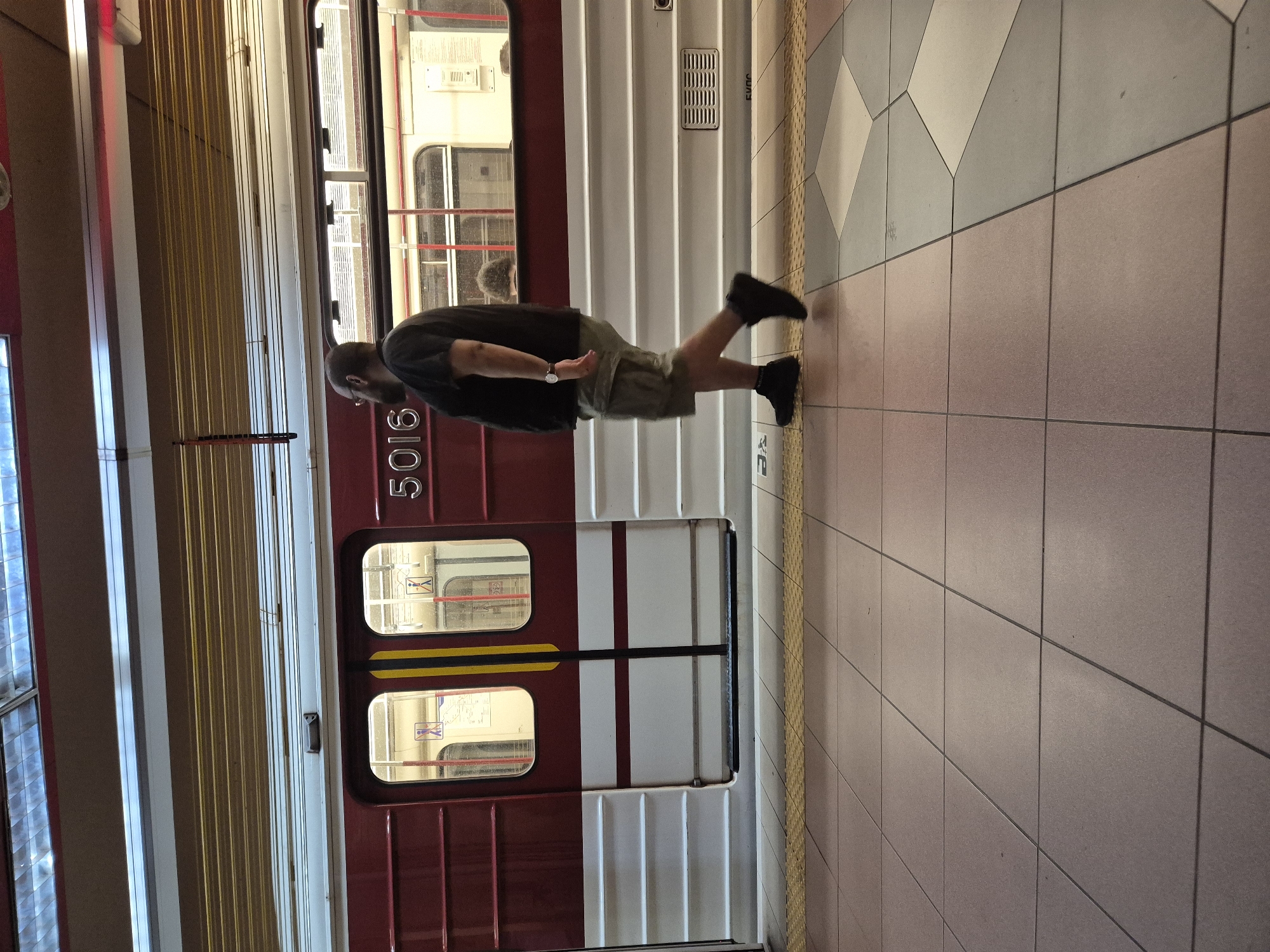 A man with dark hair and a beard, wearing a black t-shirt and khaki shorts, walks along a tiled subway platform. He is in profile, facing left. Behind him is a red and white subway train with the number "5016" visible on its side. The platform has yellow tactile paving at the edge. The train appears to be at a station in Sofia, Bulgaria.