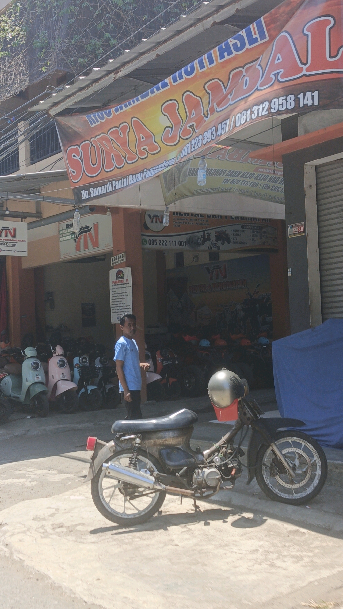 The image shows a street scene in Desa Pagandaran, Indonesia. In the foreground, a weathered motorcycle is parked on a paved surface. Behind it, a man in a blue t-shirt and dark pants stands to the left, facing towards the viewer and slightly to the right. A banner with large, orange lettering reading "SUKYA JAMAL" is prominently displayed above a building. The building appears to be a rental business, with signs indicating "RENTAL DAN PENGINAPAN" (Rental and Lodging) and various vehicle types such as ATVs and mini-cross. Several scooters and ATVs are parked under a covered area of the building. The environment suggests a sunny day, with shadows cast on the ground.