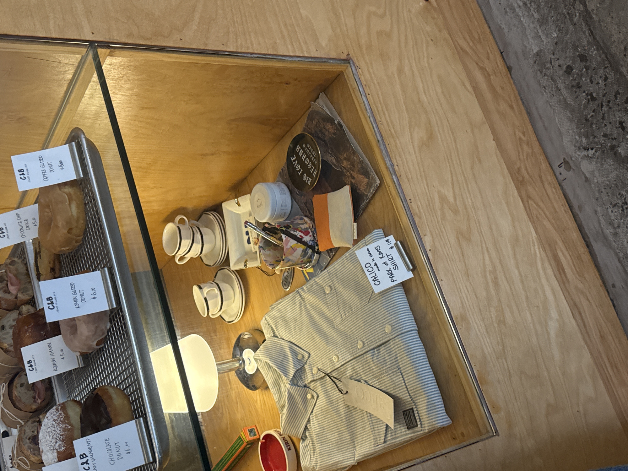 Here is a forensic description of the image:
The image shows a display case in a business located in the East Village neighborhood of New York City, United States of America. The case contains food items on the left side of the image, specifically a tray of donuts labeled with "C&B EAST VILLAGE, NY," along with various price tags for items like "CHOCOLATE DONUT $6.00," "KOUIGN AMANN $5.50," "LEMON GLAZED DONUT $6.00," and "CHOODLATE CHIP COOKIE $5.00." To the right of the food is a display of other items, including teacups, a cream jar, an art supply, a "BIG LOVE RECORDS HARAJUKU TOKYO" item, and a folded light blue and white striped shirt marked as "CALICO SHIRT PARK at KIMS, made in Korea, $119."
