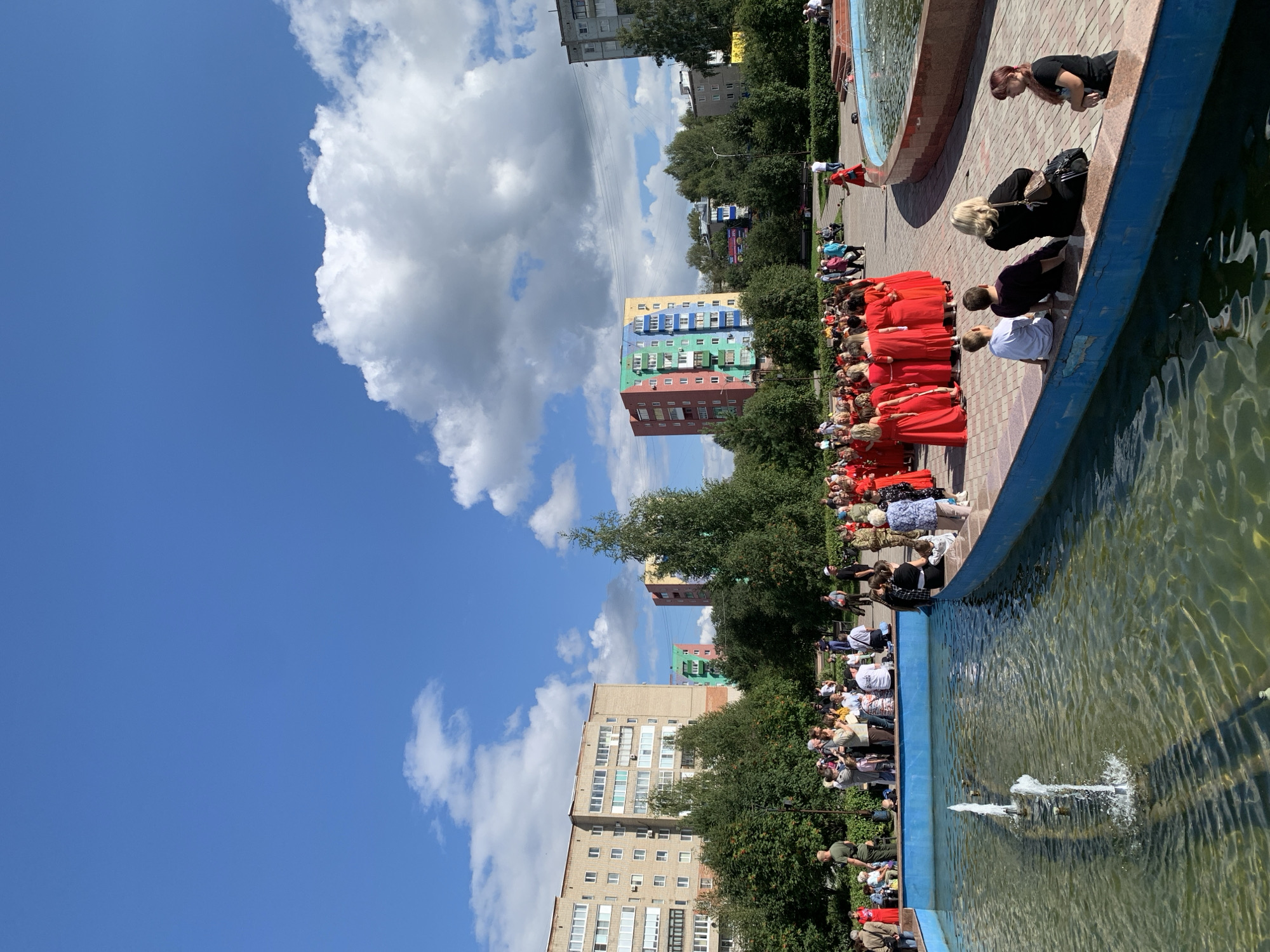 The image depicts an outdoor scene with a group of people gathered near a fountain in a public space, possibly a park or square. The setting appears to be urban, with several apartment buildings visible in the background. Some of the buildings have colorful facades, suggesting a potentially vibrant residential area. Lush green trees line the area, contributing to a park-like atmosphere.
In the foreground, a large fountain with clear water and visible water jets is a prominent feature. People are congregated around the fountain and in the paved area adjacent to it. A significant portion of the group, particularly on the left side of the image, consists of individuals dressed in matching bright red outfits, suggesting they might be participating in an event, performance, or festival. Other individuals are dressed in casual attire. Some people are standing, while others are seated on the edge of the fountain or on benches.
The sky is blue with scattered white clouds, indicating fair weather. The overall scene suggests a community gathering or public event taking place on a sunny day. The specific location in the city of Prokopyevsk, Russia, is confirmed by the context.