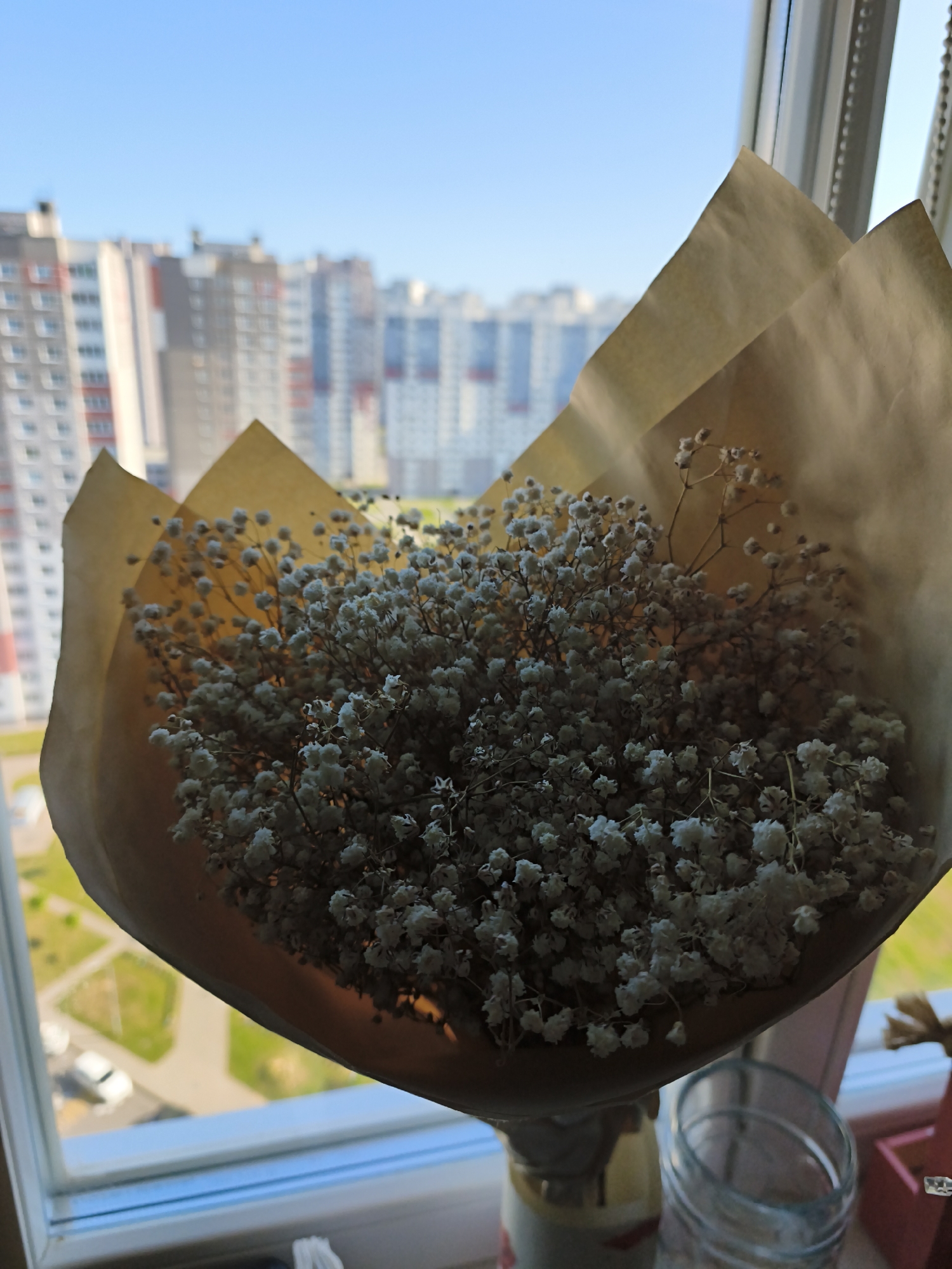 A close-up, slightly angled shot shows a bouquet of small white flowers, likely baby's breath, wrapped in brown paper, positioned in front of a window. The flowers are densely packed and appear to be dried. Beyond the flowers, through the window, a blurred cityscape of tall, multi-story buildings under a clear blue sky is visible. The buildings are a mix of beige, white, and grey, with some red accents. In the foreground, on what appears to be a windowsill, a clear glass jar and the top of a vase are partially visible. The overall lighting suggests an outdoor daytime scene.