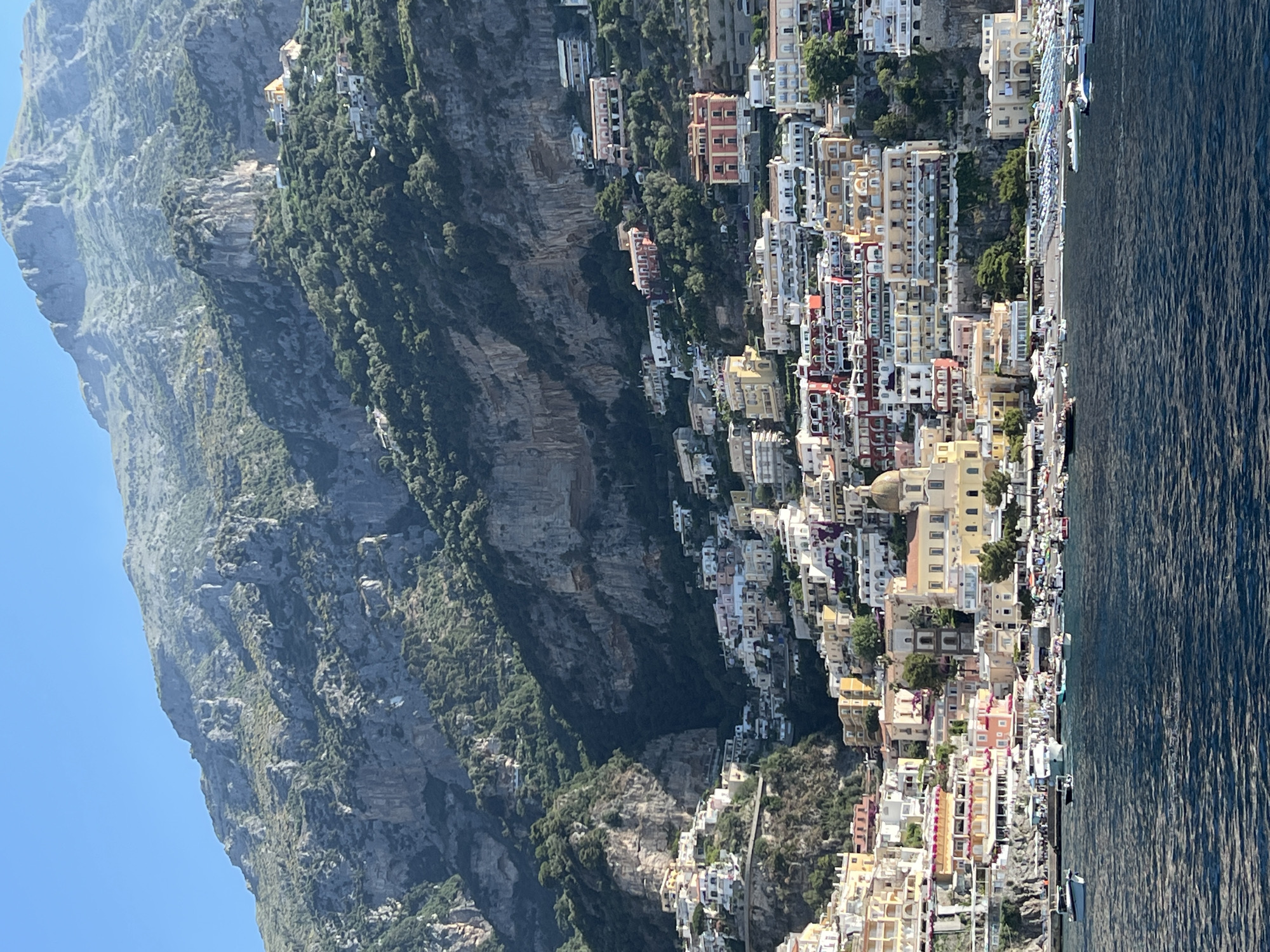 This is a photograph of the coastal town of Positano, located in Arienzo, Italy. The image captures the vibrant, colorful buildings of the town cascading down a steep mountainside towards the sea. The architecture is densely packed, with houses and structures of various sizes and shapes built into the cliff face. Many buildings are painted in pastel colors like yellow, pink, and white, with red tile roofs. A prominent church dome, possibly part of the Church of Santa Maria Assunta, is visible among the buildings.
Below the town, a shoreline is lined with a beach filled with numerous lounge chairs and umbrellas, suggesting a tourist destination. A harbor area is bustling with a variety of boats, including small motorboats and larger yachts. The water in the foreground is a deep blue, with ripples indicating a gentle sea. The background is dominated by a rugged, green mountain range, with sheer rock faces and dense vegetation. The sky above is a clear, bright blue, indicating fair weather. There are no individuals clearly identifiable or engaged in any specific actions within the frame. The scene is a picturesque view of a Mediterranean coastal town.