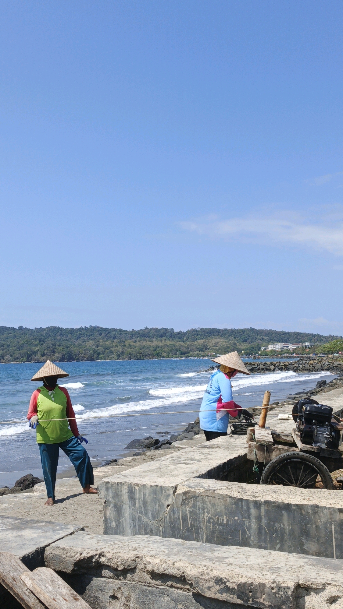 Two individuals are on a concrete embankment near a beach in Desa Pagandaran, Indonesia, under a clear blue sky. The person on the left, wearing a conical straw hat, a green long-sleeved shirt with red sleeves, blue pants, and blue gloves, stands facing the ocean. The person on the right, also wearing a conical straw hat, a blue long-sleeved shirt with pink accents, and pink gloves, is bent over, seemingly working with equipment. In the background, the ocean has moderate waves, and a lush, tree-covered hillside forms the backdrop. A small, dark-colored engine with a wheel is visible on the embankment near the person on the right. The concrete embankment shows signs of wear and tear.