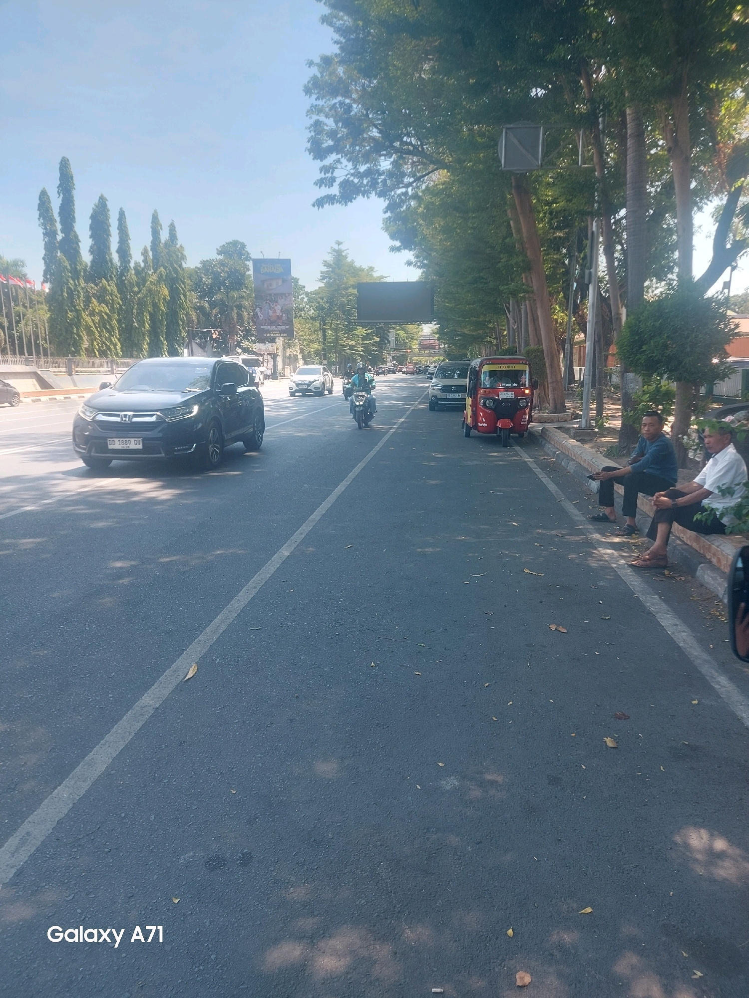 The image shows a street scene in Makassar, Indonesia, with vehicles and pedestrians present. On the left side of the road, a dark-colored Honda CRV is driving. In the background, there are tall trees and buildings, with a billboard visible. A red auto-rickshaw, a common mode of transport in Indonesia, is parked on the right side of the road. Two men are sitting on a bench on the right sidewalk, observing the traffic. The road is paved and marked with white lines. The sky is clear and blue, indicating a sunny day. The overall impression is of a typical urban environment in Makassar, with a mix of modern and traditional elements.
