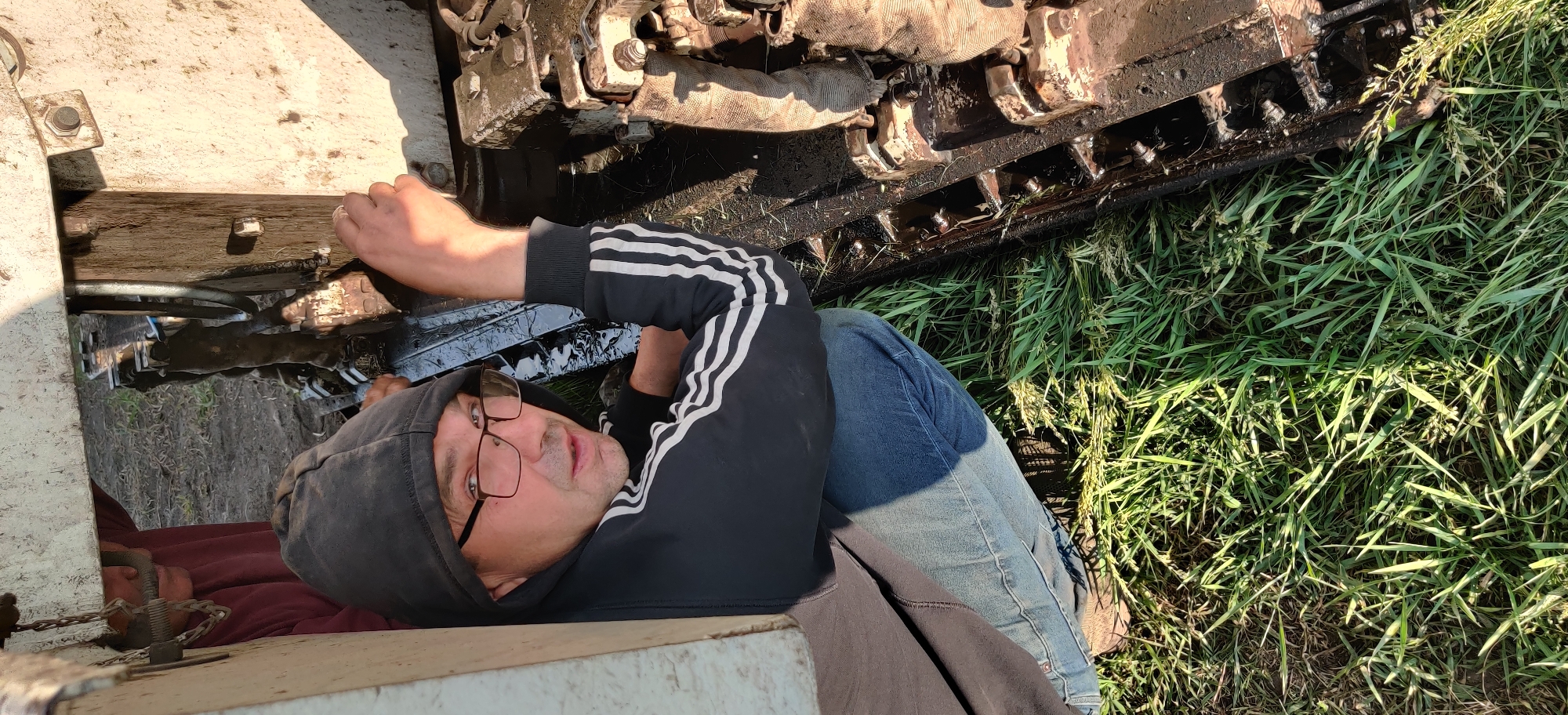 A man wearing a black hoodie with white stripes on the sleeves and blue jeans is lying on his back, appearing to work on a tracked vehicle. He is wearing glasses and looking towards the camera. The tracked vehicle is greasy and near some grass. The location is in Randava, Ukraine.