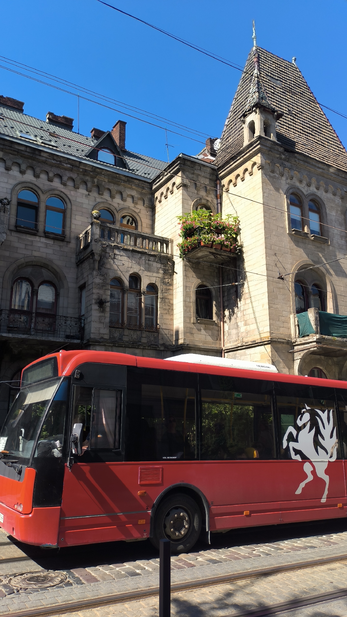 This is a medium shot, eye-level, vertical photograph of a red bus in the foreground with an ornate, historic stone building in the background under a clear blue sky. The bus is facing left, and its side is visible, featuring dark tinted windows and a white graphic of a horse on the right side. The building behind the bus is made of light-colored stone blocks and has multiple windows, balconies with decorative railings, and a steeply pitched tiled roof with a pointed turret. There are flower boxes with pink and red flowers on a balcony. The bus is on a cobblestone street with tram tracks. Overhead, thin wires are strung across the frame. The scene appears to be in the city of Lviv, Ukraine, likely in the historic center, given the architecture. There are no visible people, actions, or interactions in the scene. The lighting suggests it is daytime, with strong sunlight casting shadows.