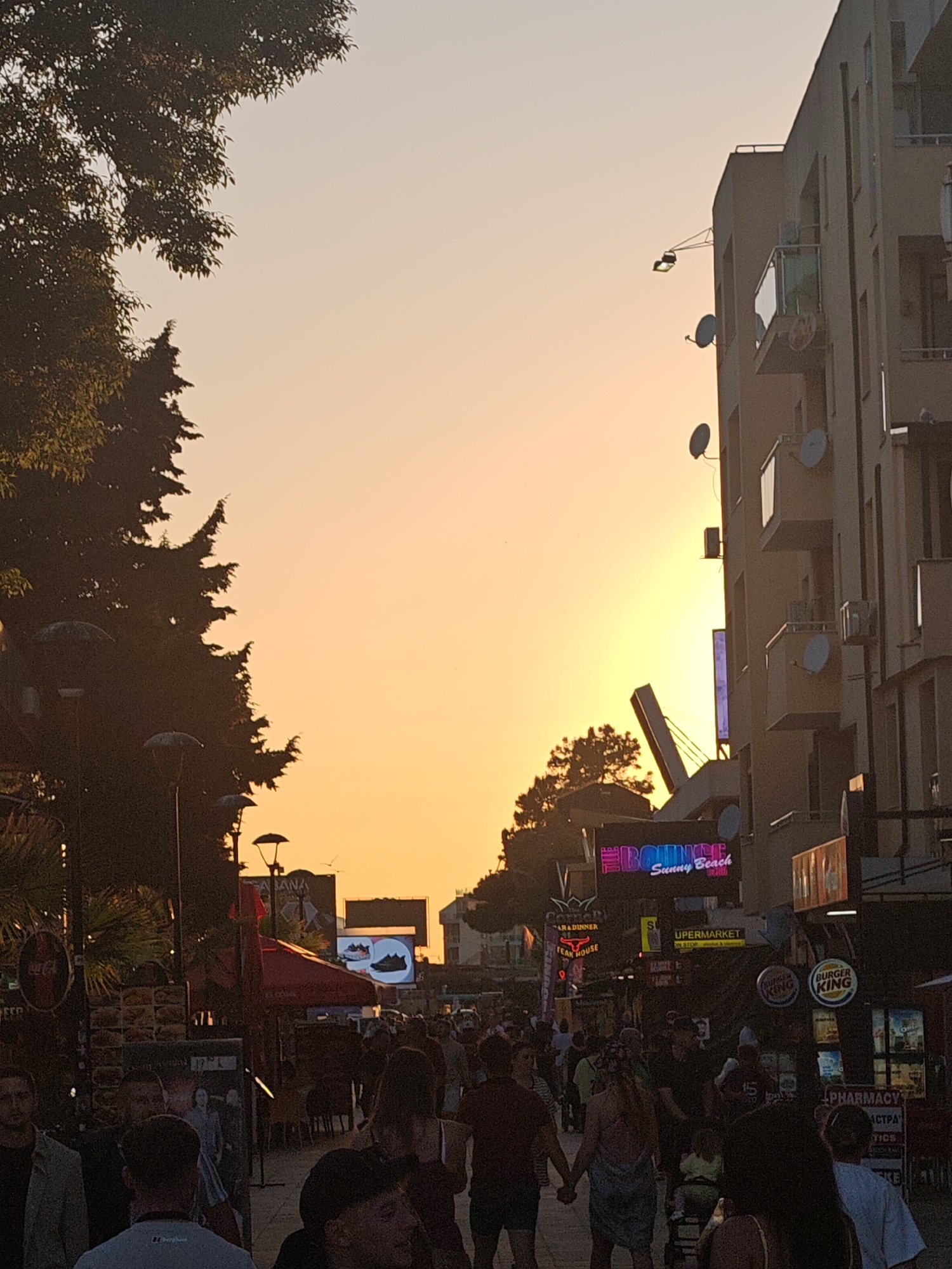 The image depicts a street scene in Sunny Beach, a resort area within the city of Nesebar, Bulgaria. A crowd of people are walking down a street lined with businesses, evident by the various signs and establishments. A large building stands to the right with satellite dishes attached to its exterior. Street lights are visible on the left side of the street, and trees frame the edge of the frame. A setting sun is visible in the distance, casting a warm glow over the scene. Signs for various businesses are visible including Burger King, a steak house, supermarket, and a pharmacy.