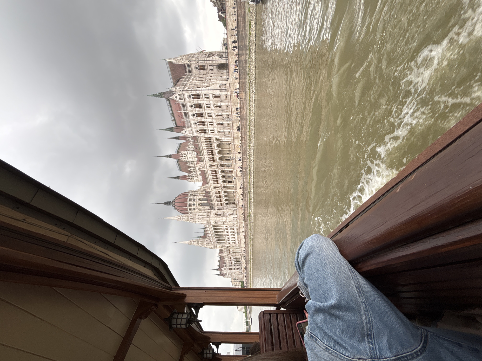 The image shows a person sitting on a boat, with their legs visible in the foreground. The boat has wooden paneling and railing. In the background, the Hungarian Parliament Building is visible on the bank of the Danube River in Budapest, Hungary. The sky is overcast. The river water appears choppy due to the boat's movement.