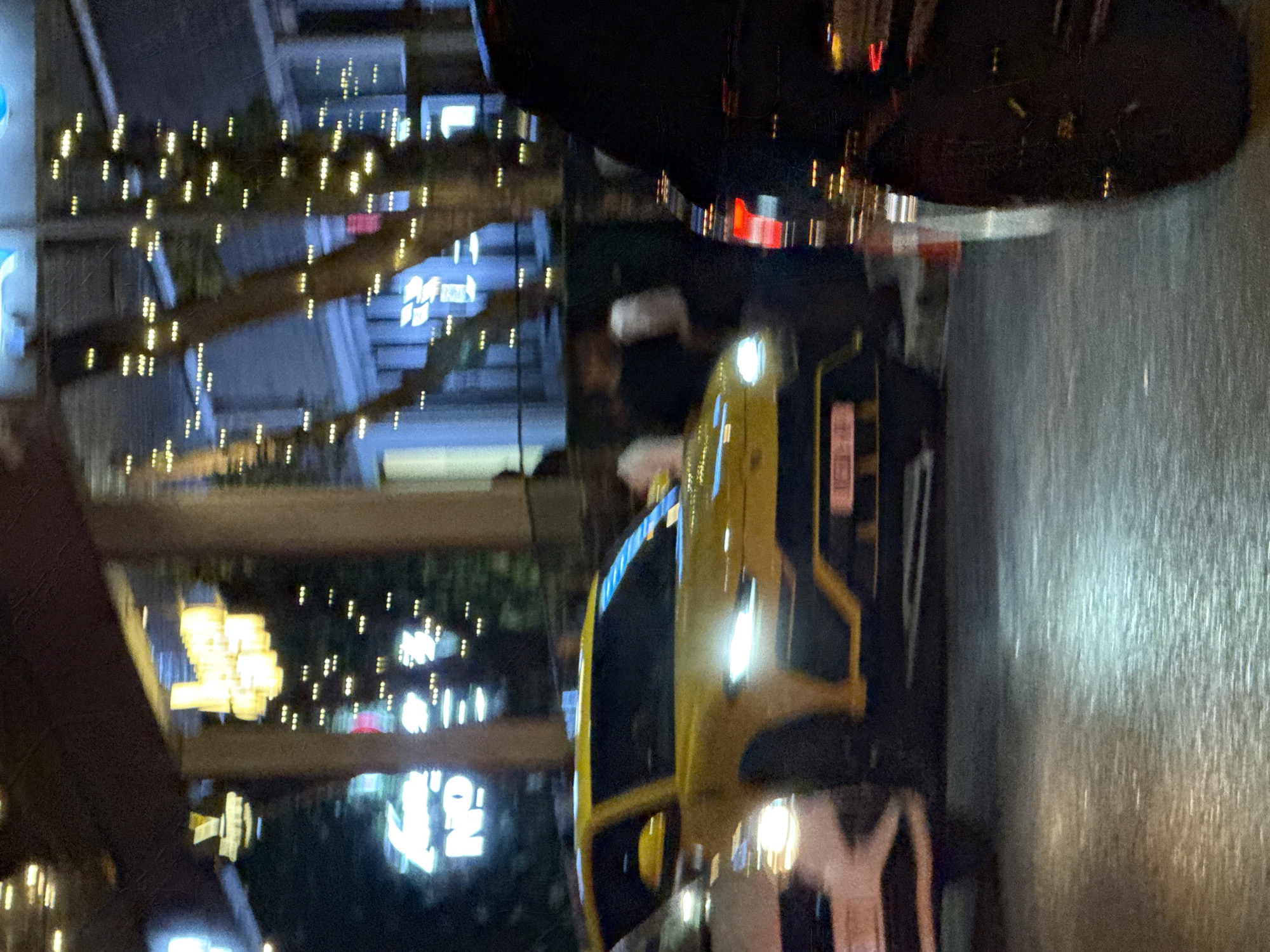 The image depicts a night scene on a street in Cape Town, South Africa, with a yellow car in the foreground and various blurred lights suggesting a city environment. The car's headlights are on, and the street appears wet, possibly after rain. There are streetlights and building lights in the background, some adorned with decorative lights, indicative of a commercial or urban area. People are vaguely visible near the car, but their actions and identities are unclear. A black vehicle is above the car.
