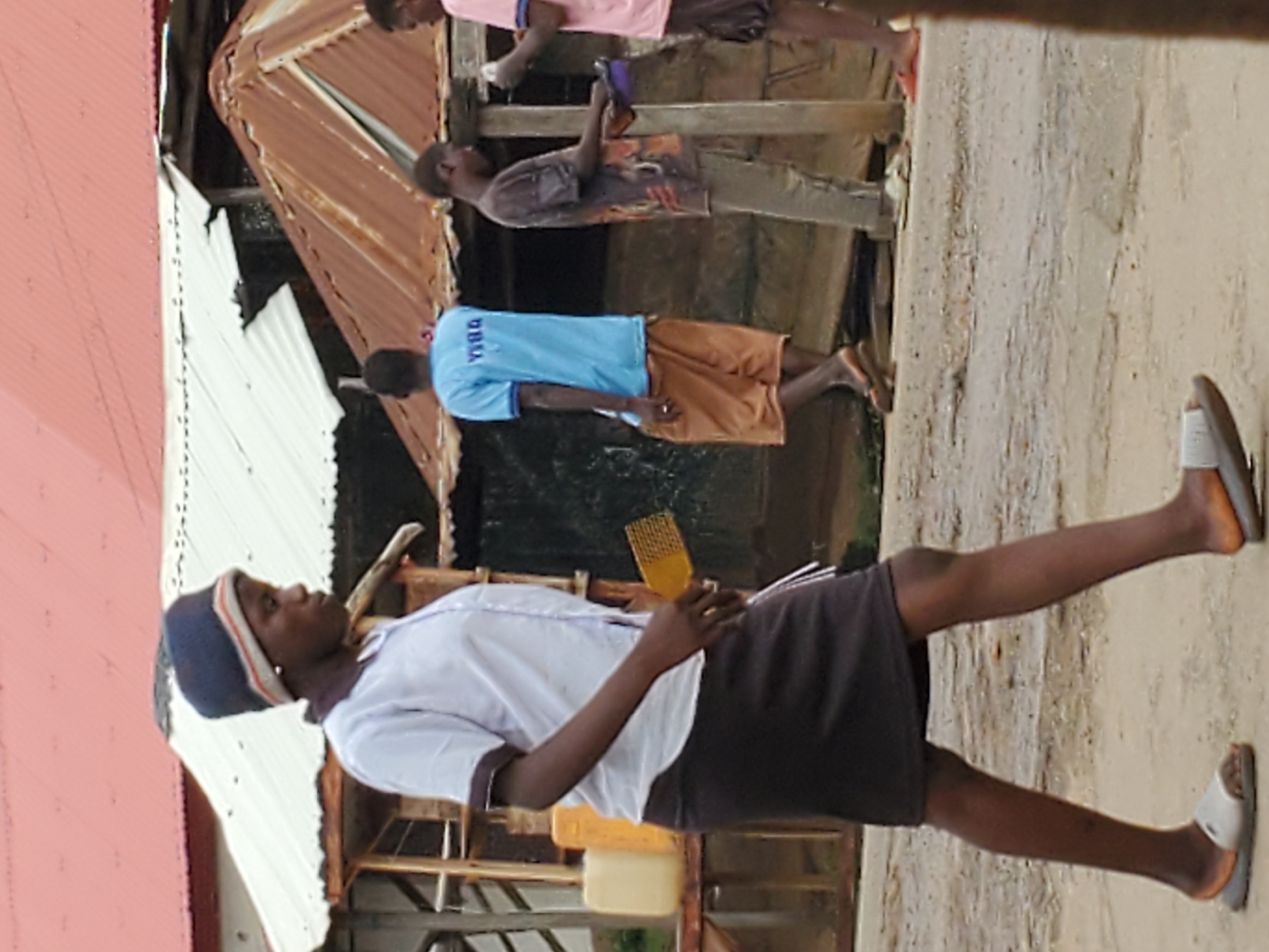 In Mararaba, Nigeria, a woman wearing a navy blue beanie, a white short-sleeved shirt, and a black skirt stands on a concrete surface. She holds a yellow comb in her right hand and a yellow container in her left. She is wearing grey sandals on her feet. Behind her, a wooden structure with a white corrugated iron roof is visible. Further in the background, three men are standing on the same concrete surface; one is wearing a blue t-shirt and brown shorts, another a brown t-shirt and brown pants, and the third a pink t-shirt and dark pants. A rusted metal roof is behind them.