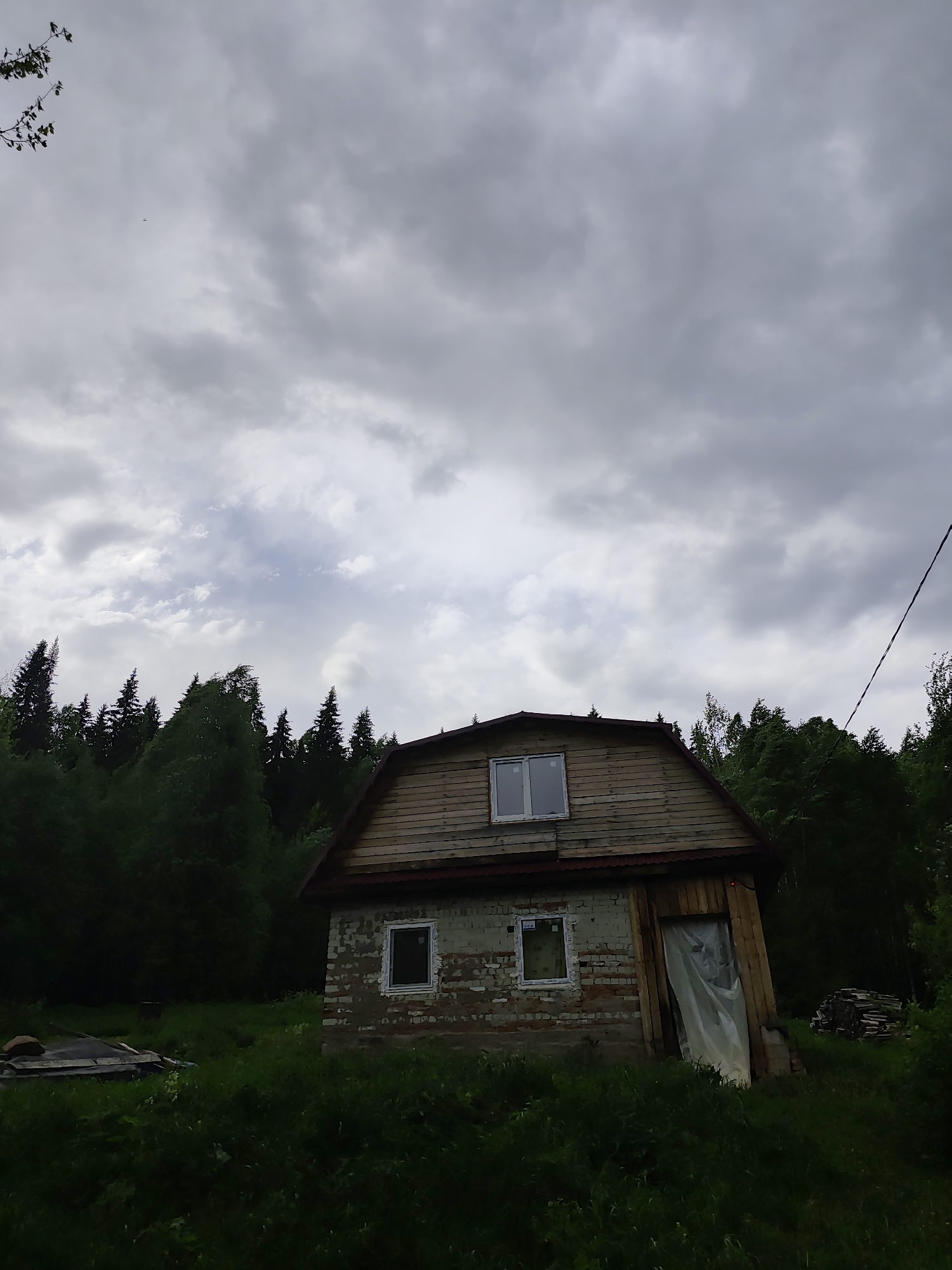 The image shows a small, partially renovated brick and wood house in an overgrown grassy area. The house has two visible windows on the lower level, framed in white, and one window on the upper level. The door is covered with a semi-transparent material. A forest surrounds the house. The sky is overcast with heavy clouds. A power line is visible on the right side of the image. Some building materials are piled up near the house. The image was taken in a rural-seeming area. This location is in Vil'va, Russia.