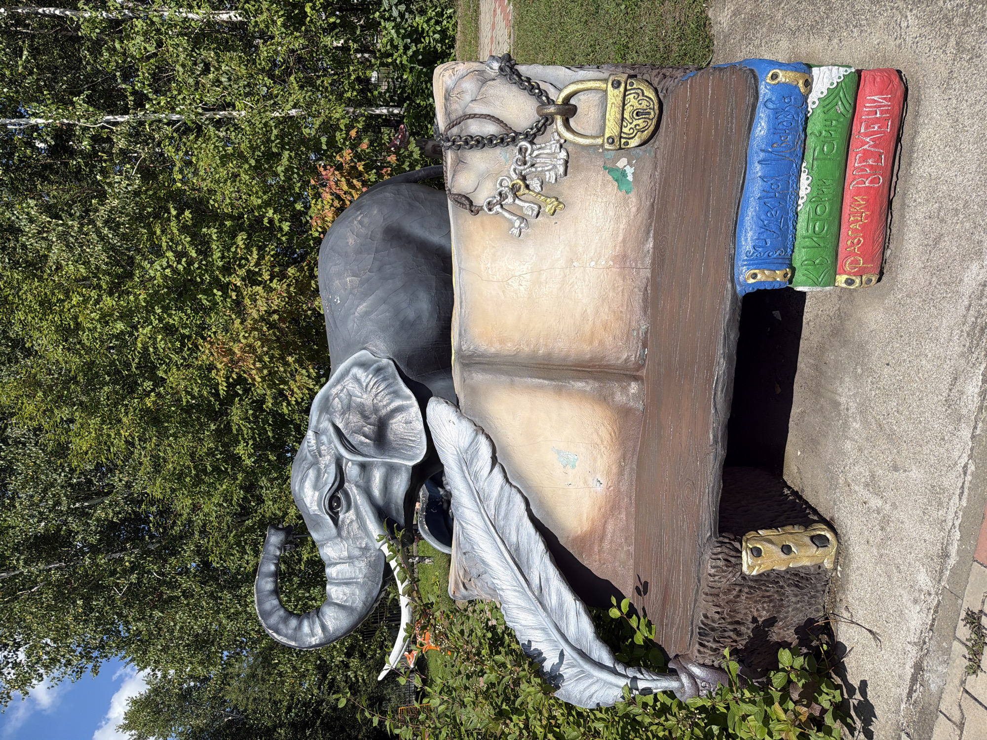 In the city of Ivanovka, Russia, a whimsical concrete sculpture depicts an elephant perched atop a large, open book. The elephant is gray with prominent tusks and its trunk is raised. Attached to the book are several keys and a golden padlock. A large feather quill rests on the book's pages. The base of the sculpture is fashioned to resemble a stack of three colorful books, with text in Russian written on their spines. The sculpture is situated outdoors, with lush green trees and foliage forming the background, suggesting a park or garden setting. The ground around the sculpture is paved with concrete and brick.