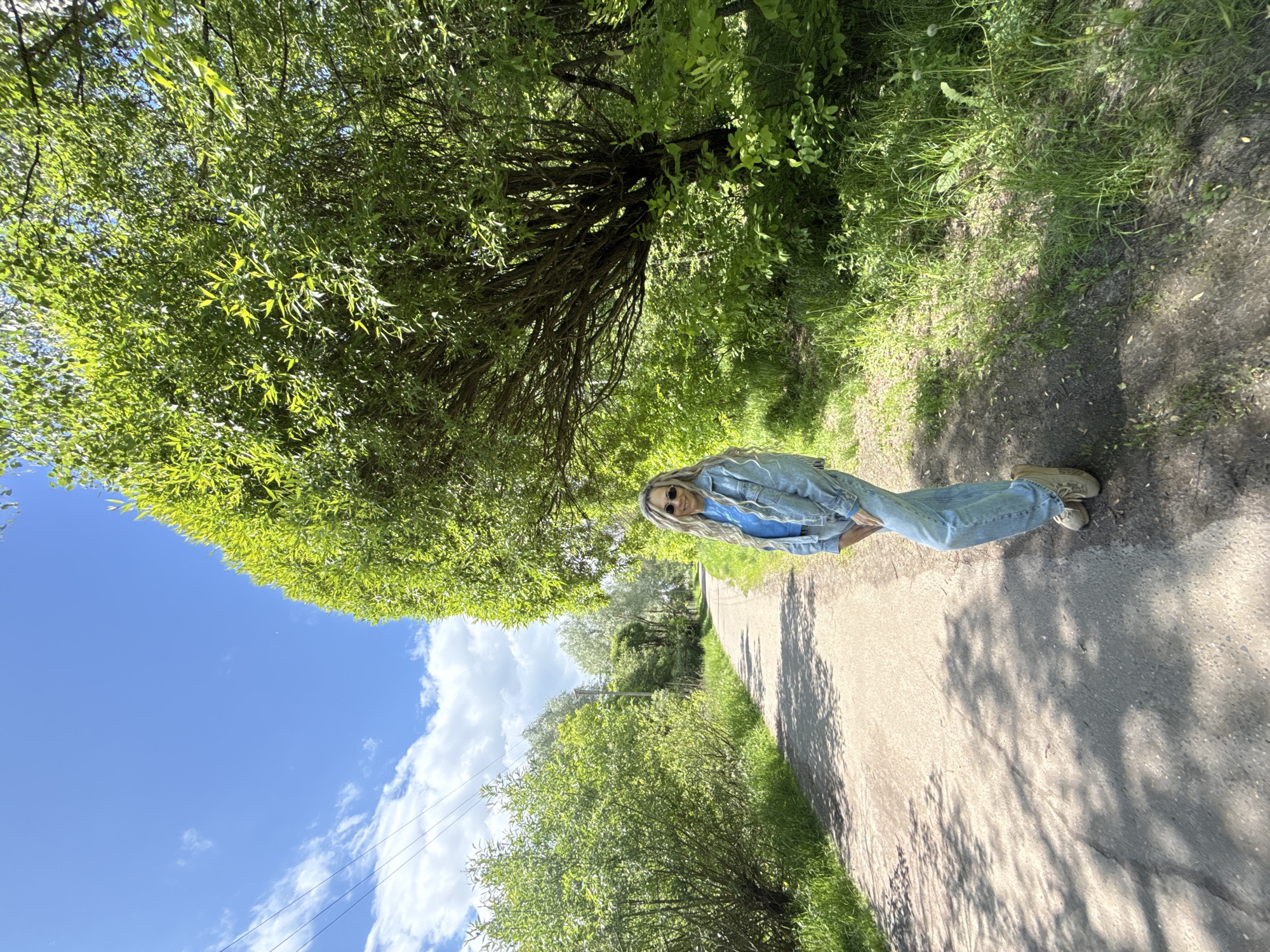 A medium shot shows a blonde-haired, fair-skinned woman with sunglasses, wearing a blue shirt and denim jacket with jeans, standing on a dirt path in a natural setting, possibly a rural area, in Шебунино, Russia. The path stretches away from the viewer, framed by green vegetation. Above, a dense tree canopy partially covers a clear blue sky with scattered white clouds. Overhead power lines are visible in the lower-left. The photo is rotated, with the bottom of the image appearing as the right side.