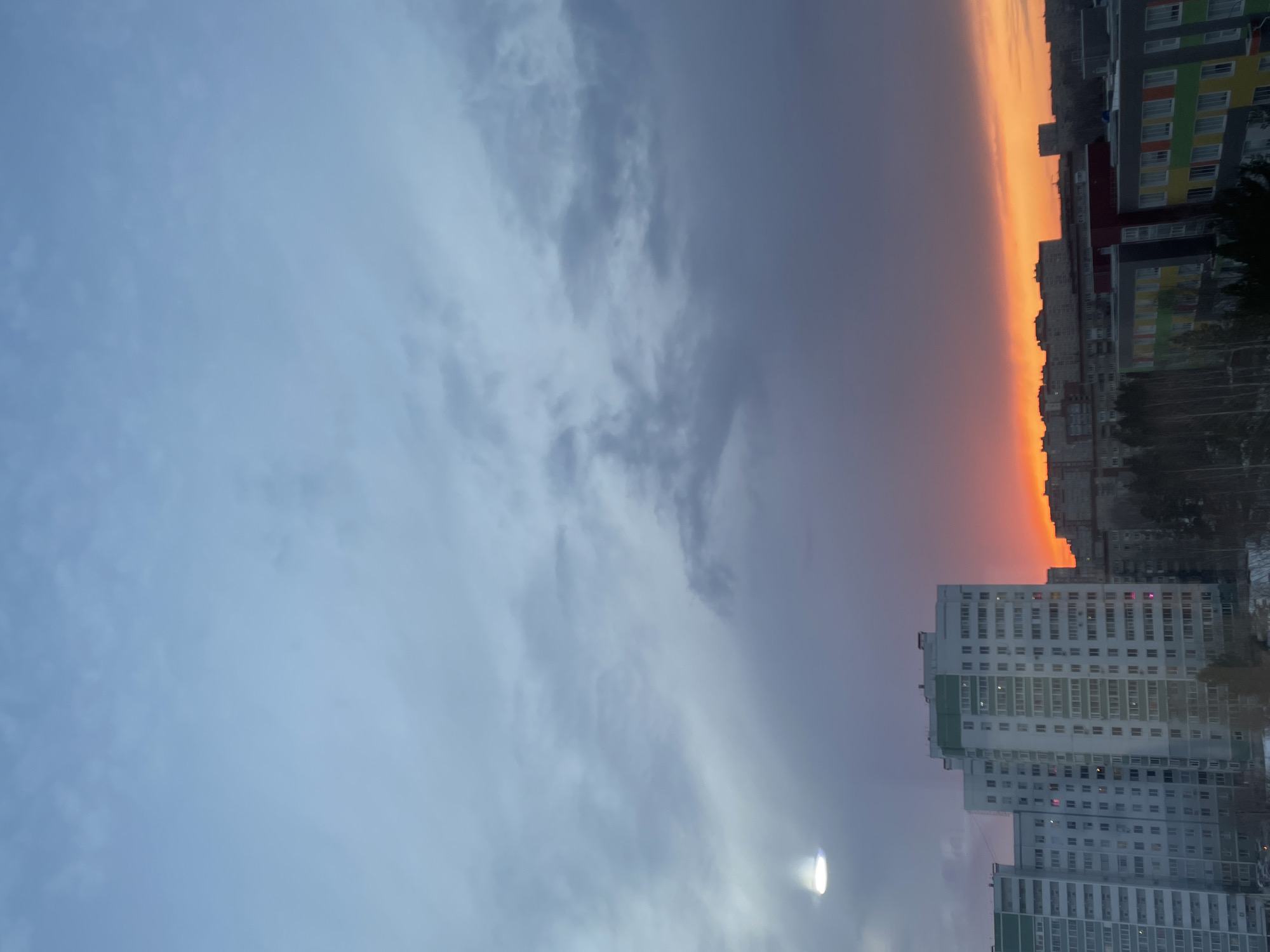 The image shows a vertical view of a cityscape at twilight, likely in Izhevsk, Russia, based on the architecture. The left side of the frame is dominated by a cloudy sky ranging from light blue to dark gray. A bright, out-of-focus light source is visible in the lower left. The right side of the frame features a row of buildings. At the bottom right, there is a large, white apartment building with multiple windows and green accents. Adjacent to it and further up the frame is another row of buildings, including one with colorful panels. Above that, the horizon is marked by a vibrant orange glow of the setting sun. The overall composition is split almost in half between the sky and the urban landscape, capturing a vivid contrast between natural and man-made elements.