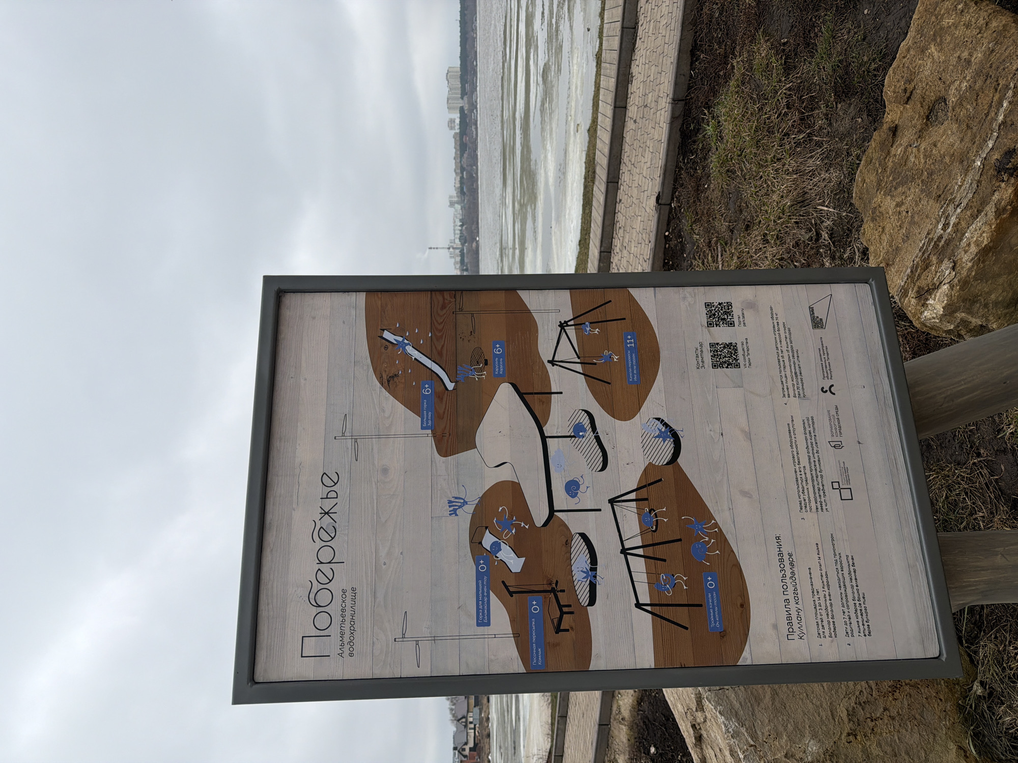 The image shows an informational sign located in the city of Almetyevsk, Russia. The sign provides information about the playground, including the rules of use and contact information. The sign is attached to wooden posts and is positioned near a paved path and a body of water. The background includes a view of a cloudy sky and buildings in the distance. A large rock is visible near the sign.