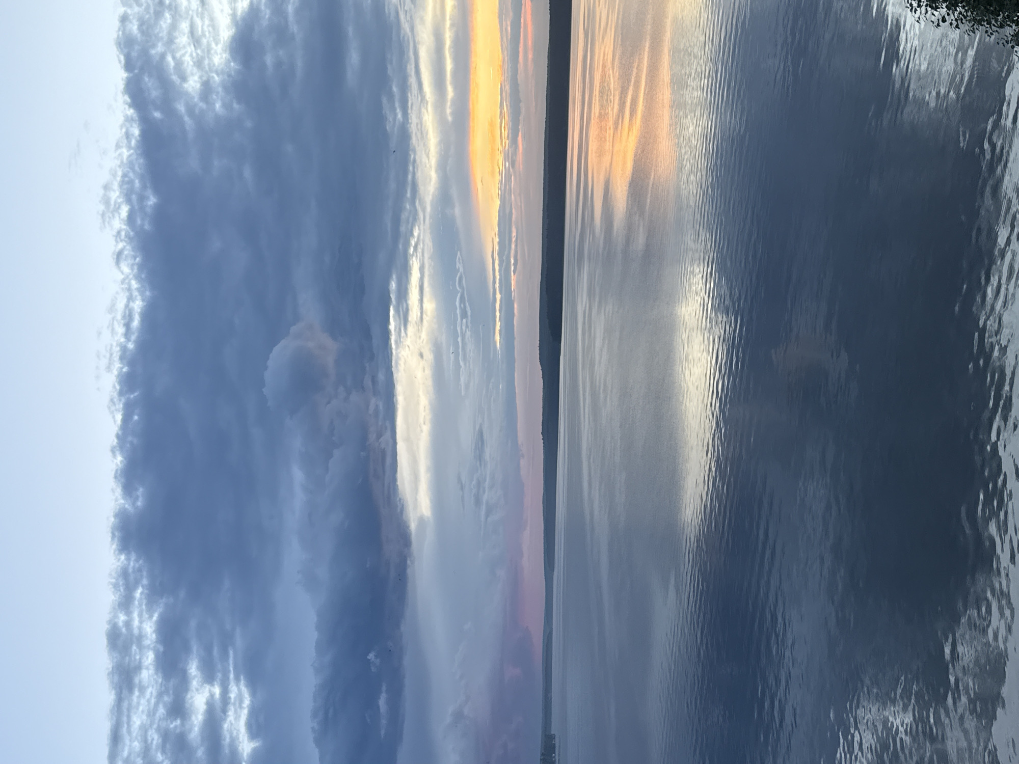 This is a vertical, landscape photograph taken from a slightly elevated perspective, overlooking a body of water that appears to be a lake or large river. The sky is filled with dramatic clouds, with a clear patch of bright orange and yellow sunlight breaking through towards the horizon, suggesting either sunrise or sunset. The light from the sun is reflected on the rippling surface of the water, creating a shimmering effect.
On the left side of the image, dark, heavy clouds dominate the sky, creating a stark contrast with the bright colors of the sunset. The water on the right side of the frame is calm and reflective, mirroring the sky above. A distant shoreline is visible as a dark, silhouetted line against the brightly lit sky.
There are no visible people or objects of note in the foreground. The overall mood is serene and tranquil, with a sense of natural beauty. The time of day is clearly either dawn or dusk due to the intense colors in the sky and their reflection on the water. The location context of Votkinsk, Russia, suggests this is likely a scene from one of the many waterways in the region. There is no visible text within the image.