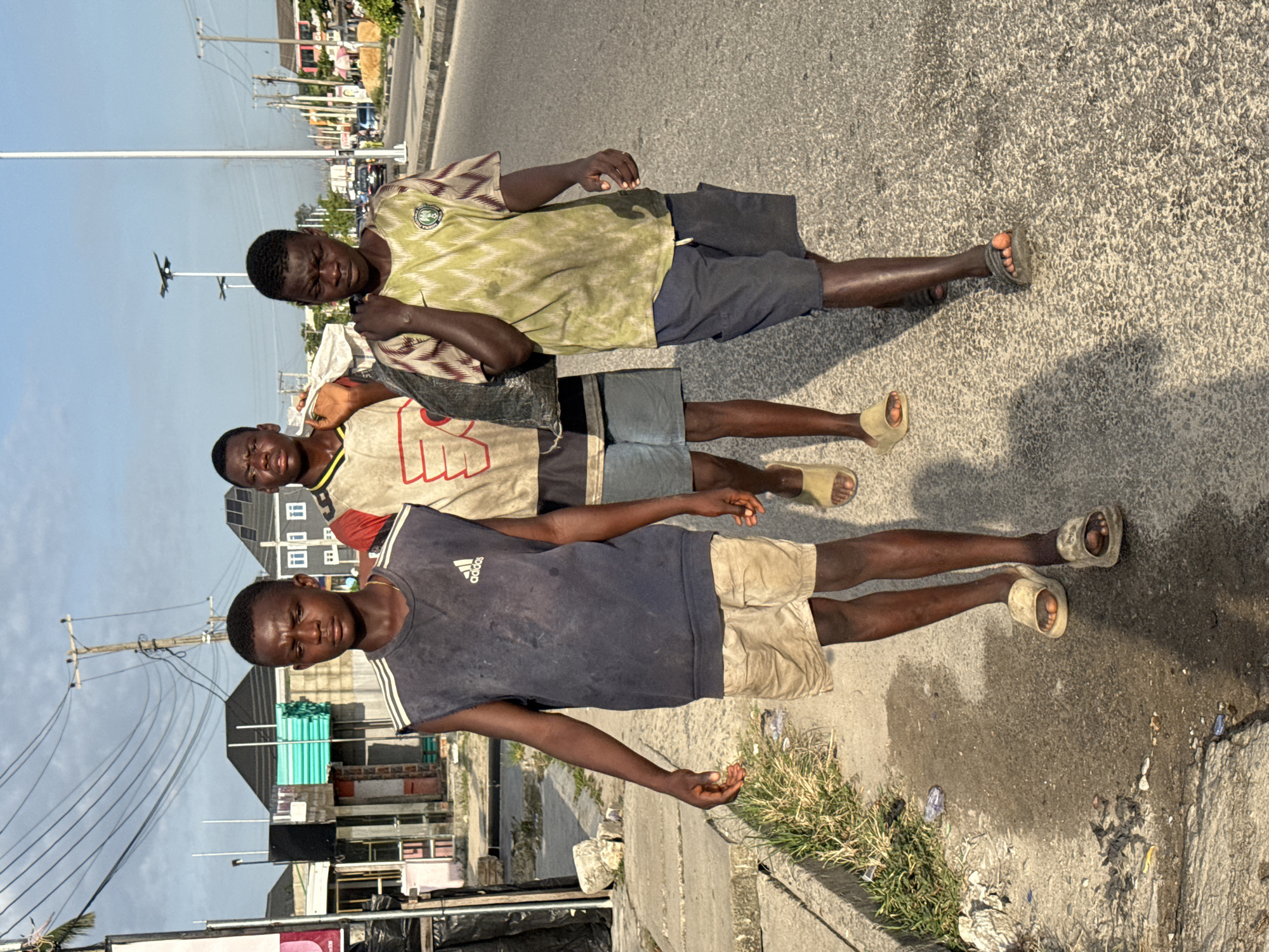 Three young men stand on a paved road in Ado, Nigeria. The man closest to the viewer wears a dark blue Adidas tank top and khaki shorts, while the others wear t-shirts and shorts. All three wear sandals. The street has litter along the edge. Buildings and power lines are visible in the background.