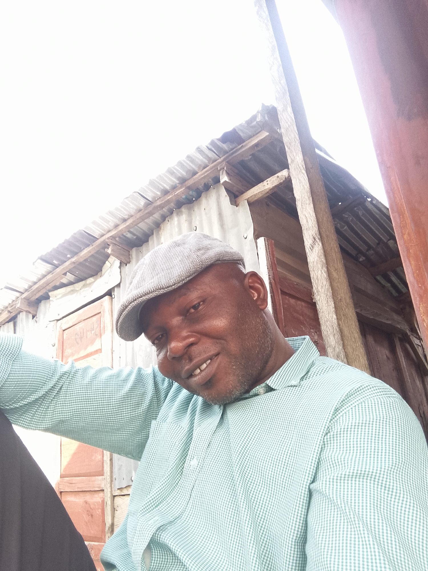 A man is posing for a selfie in an outdoor setting in the city of Unknown, Unknown. The man is wearing a gray flat cap and a light green and white checkered collared shirt. Behind him is a building constructed of corrugated metal and wooden planks. The siding of the building appears to be red/brown, and there is a wooden post situated along the right side. The sky is visible in the background, but it is heavily overexposed due to the bright sunlight.