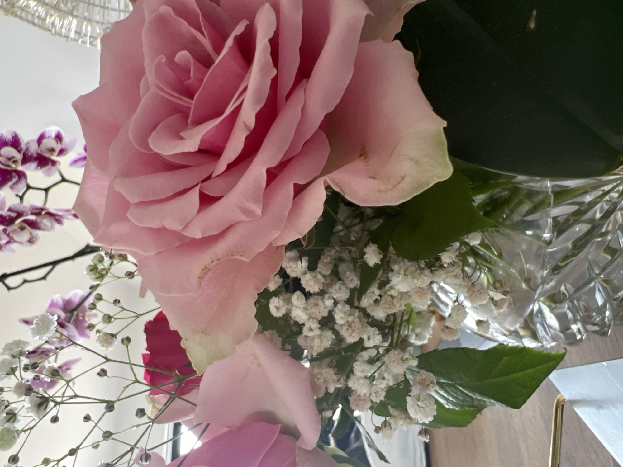 A close-up shot shows a bouquet of pink roses and white baby's breath flowers in a clear, faceted glass vase. The flowers are arranged in a way that the large pink rose is the most prominent subject, with its petals unfurling towards the viewer. In the background, blurred purple orchids are visible. The setting appears to be indoors, possibly on a wooden surface, with soft lighting. There are no people or animals present in the image. The image does not provide any information that can be used to determine its location in Bunbeg, Ireland.