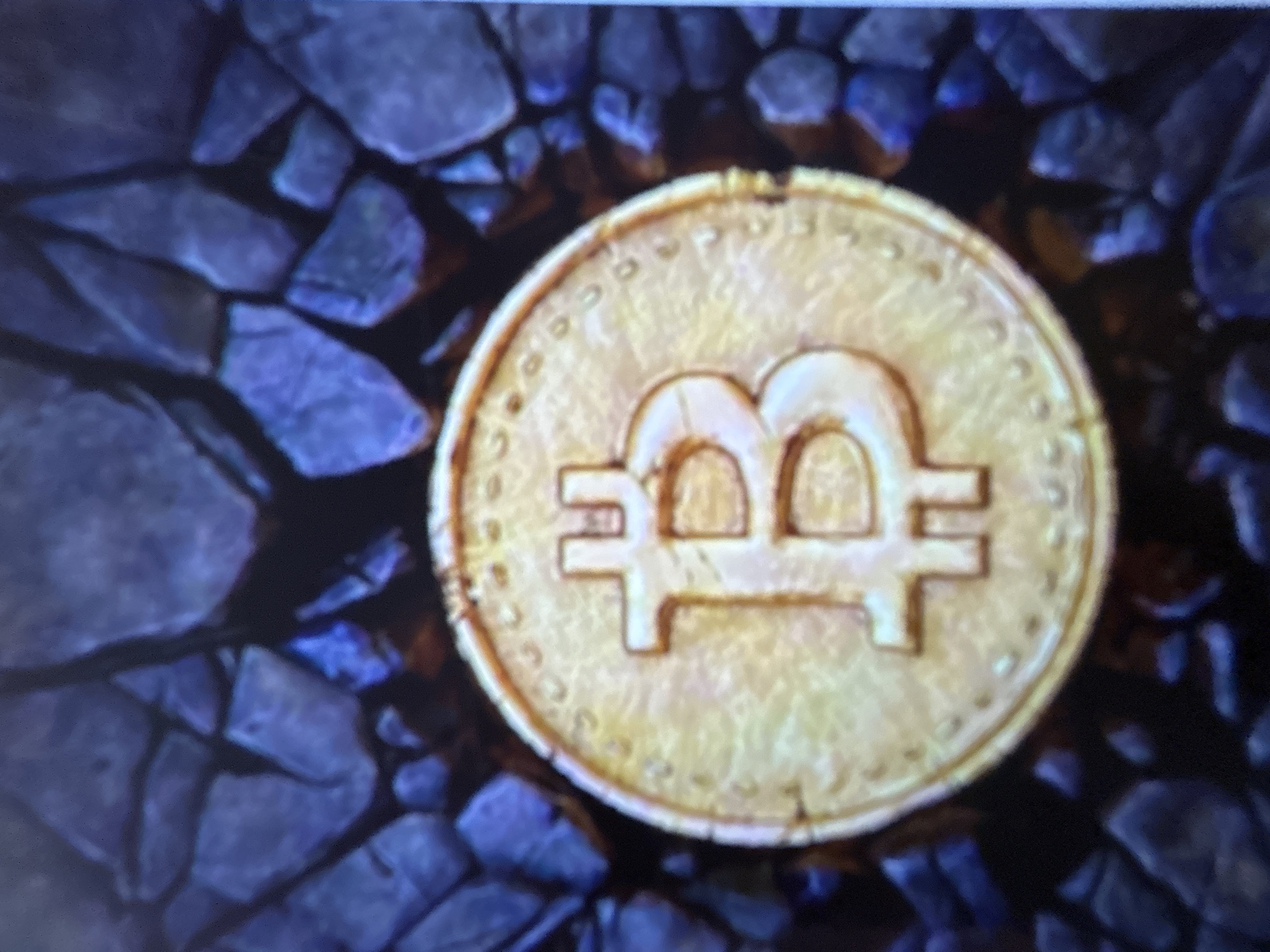 A close-up, vertical, eye-level shot displays a golden Bitcoin coin positioned centrally on a dark, textured surface resembling rough, dark blue rocks or stones. The Bitcoin coin is the primary subject, featuring a prominent, raised symbol of the letter "B" with vertical lines extending from the top and bottom, indicative of the Bitcoin logo. The coin's surface appears slightly worn and textured, with small, circular indentations along its rim. The background consists of an arrangement of dark, angular, rock-like elements that create a sense of depth and shadow. The lighting appears to be dim, with some highlights on the coin and a subtle glow emanating from some of the background stones, suggesting an artificial light source. There are no people or any discernible signs of a specific location within the city of Gelendzhik, Russia, present in the image.