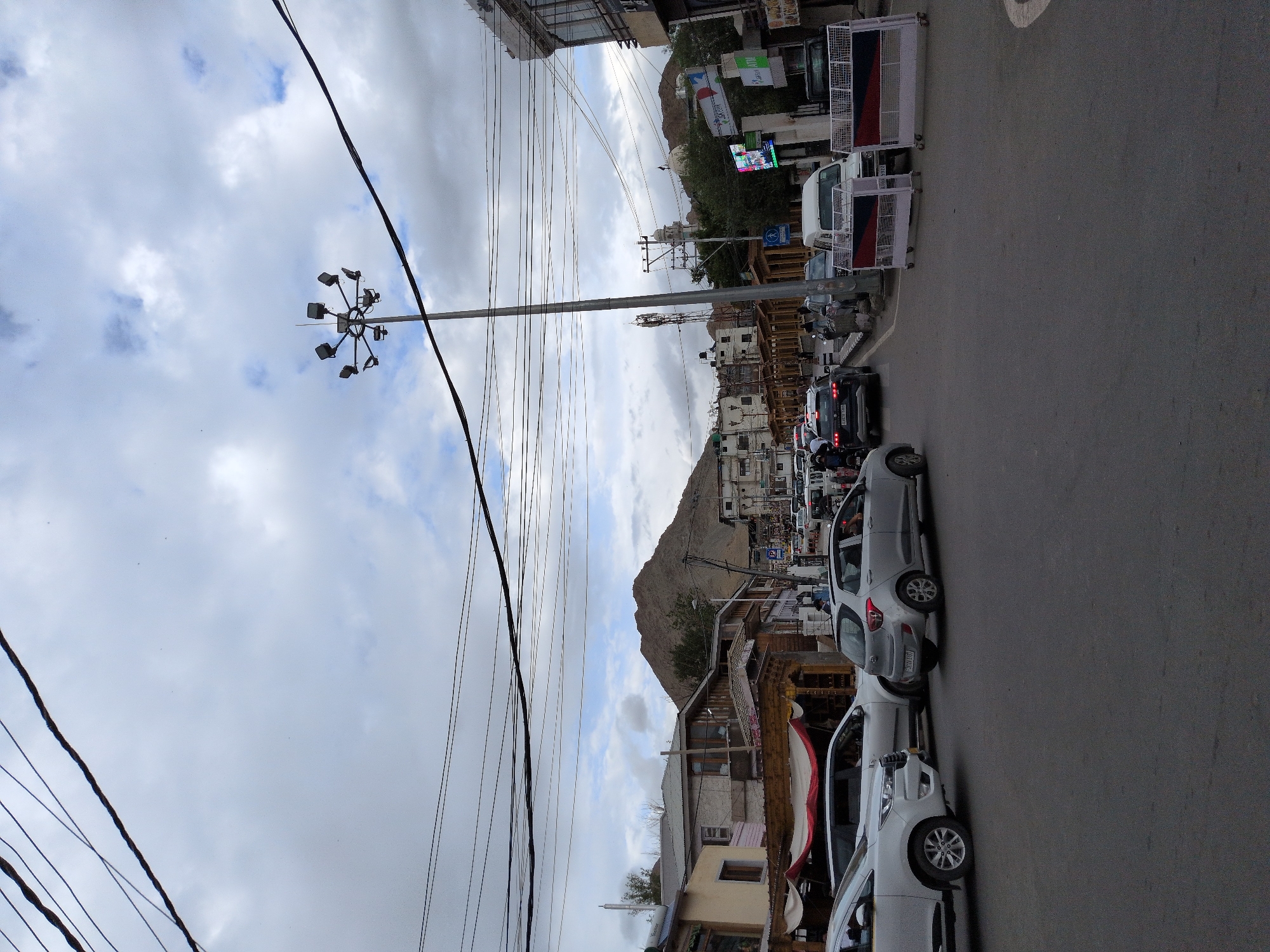This is a street-level, eye-level, medium shot of a traffic scene in Leh, India, under a cloudy sky. The foreground shows an asphalt road with several cars. In the middle ground, there is a street with buildings lining both sides and a mountain in the background. A tall light pole with multiple lamps stands near the center of the image, and several power lines crisscross the sky. A few people can be seen walking on the sidewalks. The overall atmosphere suggests a moderately busy day in the city.