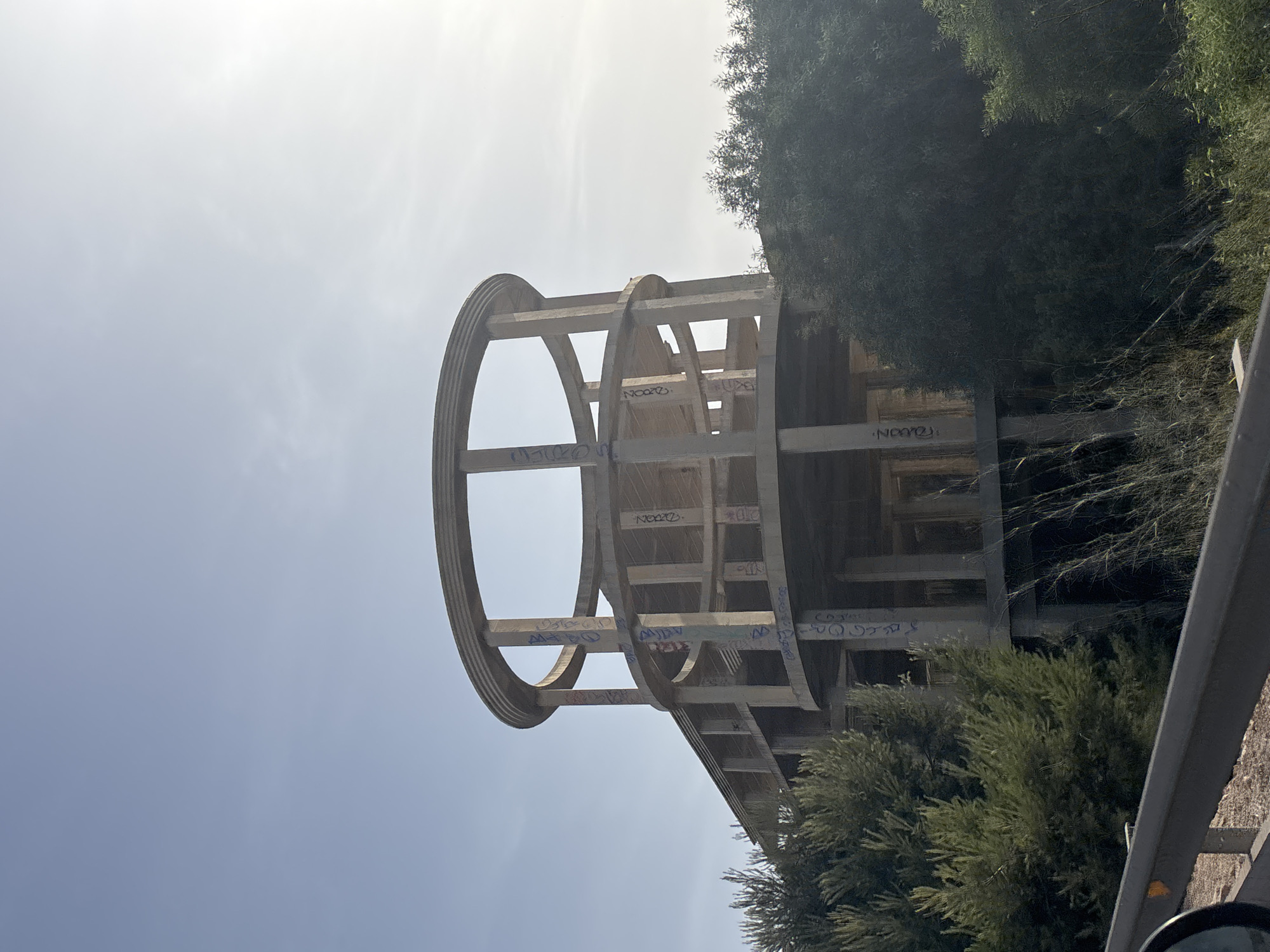 An unfinished, multi-story concrete building with a circular upper structure is surrounded by dense green foliage. The building's columns and beams are visible, and some of the concrete surfaces are marked with graffiti. The scene appears to be in an outdoor, possibly rural or semi-urban setting, with a clear sky and sunlight filtering through. A guardrail and part of a vehicle are visible in the foreground, suggesting the image was taken from a road. The location is identified as Urbanización La Cenia, Spain.