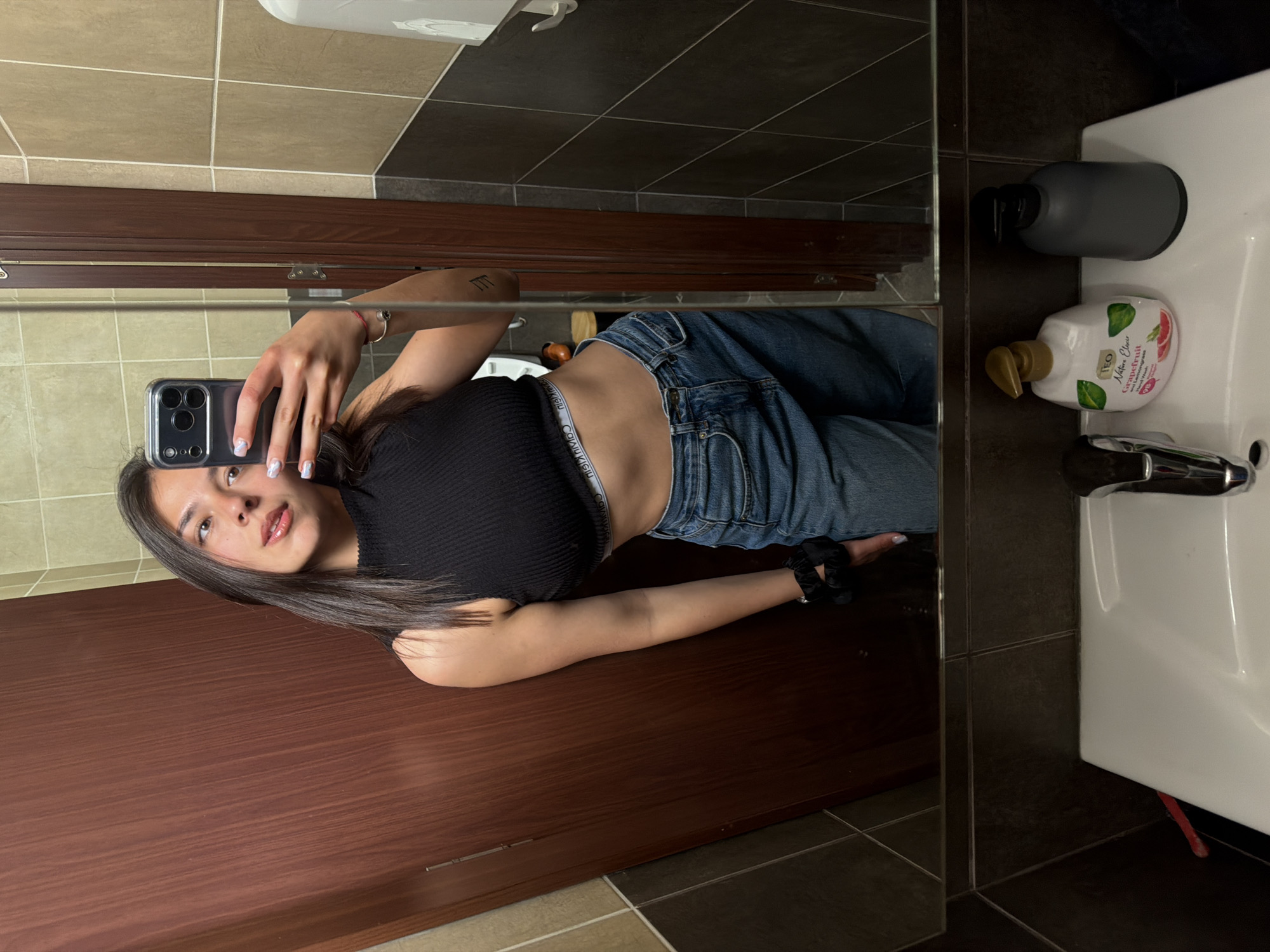 The image displays a young woman taking a mirror selfie in what appears to be a bathroom in Gabrovo, Bulgaria. She is reflected lying down or leaning against a surface, holding an iPhone with a clear case to capture her image. She has long, dark hair, light makeup, and a neutral to slightly smiling expression, looking directly into the camera.
She is dressed in a black ribbed crop top and blue denim jeans, with the waistband of her "CALVIN KLEIN" underwear visible above her jeans. A black scrunchie is on her left wrist, and she also wears a thin red string bracelet and another silver-colored bracelet on her right wrist. Her fingernails have light-colored polish. A small, unidentifiable tattoo is visible on her inner upper arm.
The bathroom features beige and dark gray square tiles on the walls and floor. A dark wooden cabinet or door is visible to the left, reflected in the mirror. On the white ceramic sink, there are two soap dispensers: one dark gray with a black pump, and one white bottle of "TEO Nature Clean Grapefruit with Pomegranate" hand soap with a gold pump. A silver faucet is also visible. A red object, possibly a toothbrush, is partially seen beneath the sink. The lighting suggests an indoor setting, likely daytime or well-lit evening.