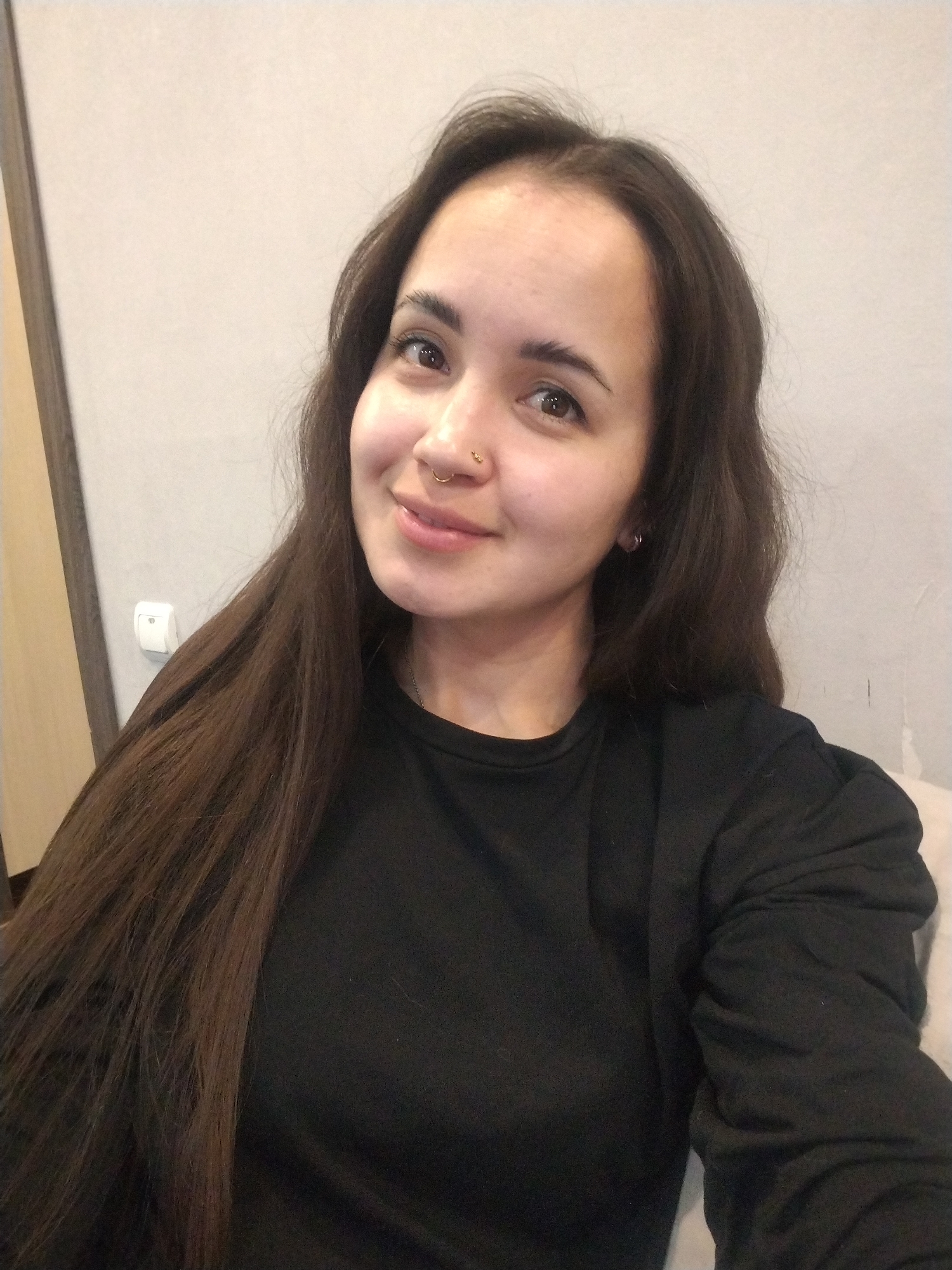 A young woman with long, dark brown hair smiles at the camera. She is wearing a black long-sleeved shirt. She has a nose piercing. The background is a light gray wall with a doorframe visible on the left. A light switch is on the wall near the doorframe. The location, Noviy, Russia, is assumed but cannot be verified.