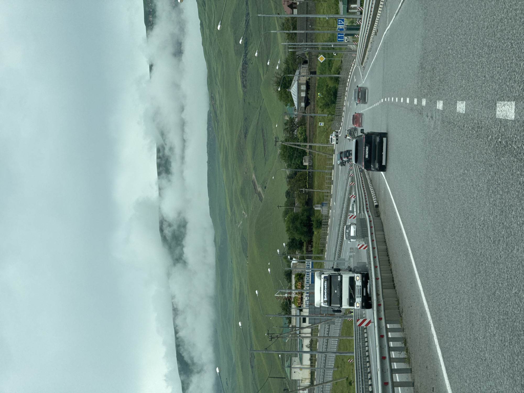 This is a vertical, eye-level, outdoor, medium shot photograph taken from a vehicle traveling on a multi-lane asphalt highway. The highway has a concrete barrier on the left and a metal guardrail on the right, both with reflective markers. The road surface is wet and appears to have standing water in some areas, suggesting recent rain.
In the foreground, a black SUV is driving in the right lane, followed by a red sedan and a dark gray sedan. In the left lane, a white semi-truck with a logo that reads "jobstratta" on its trailer is driving, preceded by a white passenger vehicle. The truck is branded with "Volvo" on the front.
The surrounding environment is a hilly, green landscape under a cloudy sky with low-lying fog or mist obscuring the upper portions of the hills. Scattered white objects, possibly sheep, are visible on the hillsides. On the left side of the highway, there are streetlights, utility poles, and buildings including what appears to be a gas station with signage in Russian. On the right side, there are more streetlights and some residential buildings. The overall impression is a scenic, somewhat overcast day on a highway in a rural or semi-rural area. The specific location is identified as Kumysh, Russia, implied by the context.