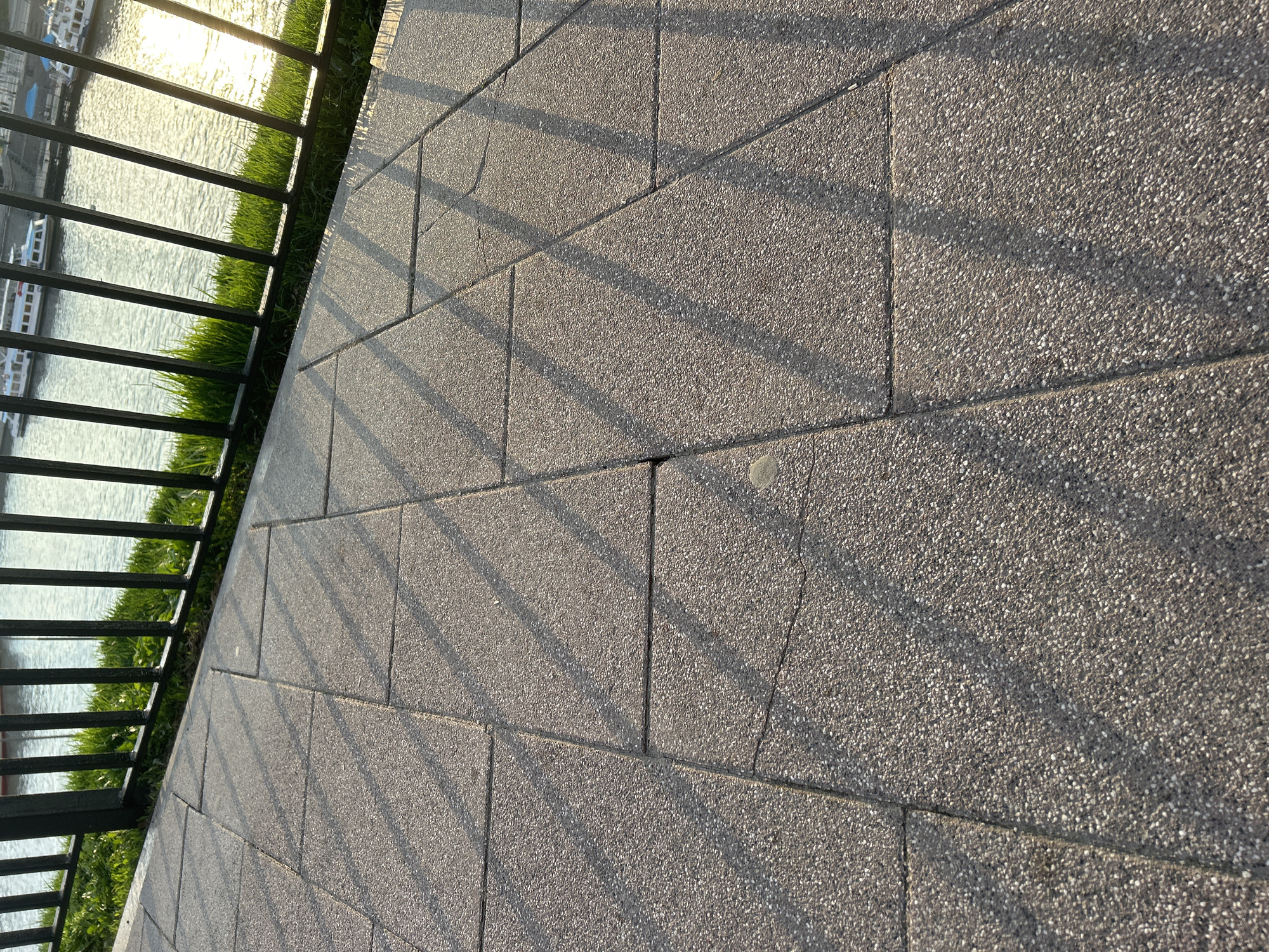The image captures an outdoor scene at what appears to be golden hour, either late afternoon or early morning, indicated by the long, diagonal shadows cast across the ground and the strong sun glare on the water. The foreground features a light grey paved walkway or patio made of rectangular, speckled pavers, running diagonally from the bottom left towards the top right. A dark, possibly black, metal fence with vertical and horizontal bars runs along the left side, separating the paved area from a strip of lush green grass.
Beyond the grass and fence is a body of water, likely a river or a large lake, reflecting the bright sunlight with a shimmering surface. Several white boats with dark accents are partially visible on the water, suggesting a marina or a busy waterway. The fence casts distinct, parallel shadows across the textured pavers, creating a pattern of light and dark bands. One paver towards the middle-right exhibits a small circular discolored spot, and some minor cracks are visible in the paving. No people or specific activities are depicted, giving the scene a serene and tranquil atmosphere. No visible text is present.