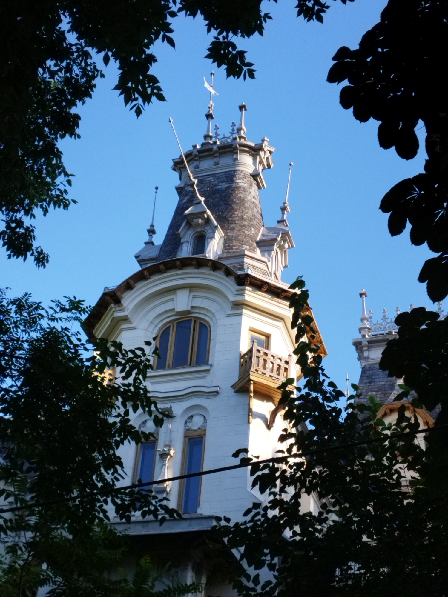 A low-angle, eye-level shot captures the top portion of a white building with a grey, conical roof and ornate architectural details. The building, located in Bucharest, Romania, features arched windows with dark blue panes and a small wooden balcony with a decorative railing. Several trees with dark green leaves partially obscure the view of the building, creating a framing effect. The sky is a clear, bright blue. The image appears to be taken during daylight hours.