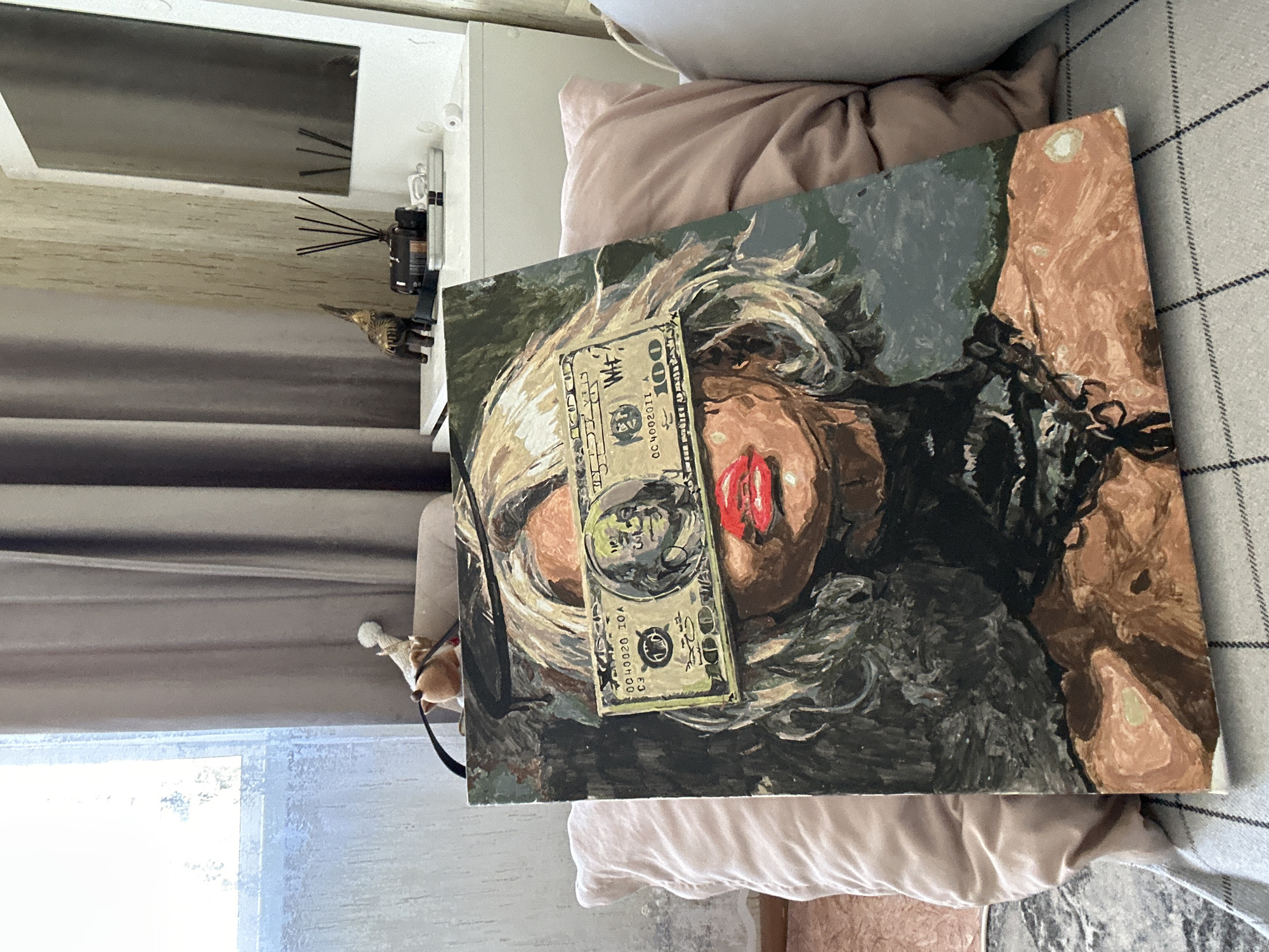 The image depicts an amateur oil painting on canvas, propped up against pillows on what appears to be a bed or sofa. The painting features a stylized, semi-abstract portrait of a person with blond hair. A dollar bill is painted over the subject's face, covering their eyes and nose, with a portion of the mouth visible. The color palette is primarily dark, with greens, browns, and blacks dominating the background and the subject's clothing. The pillows are light pink or beige. The room is illuminated by natural light from a nearby window. Other objects in the periphery include a diffuser, a small figurine, and textured curtains. There is no discernible information to indicate a specific location in the city of Высокий, Russia.