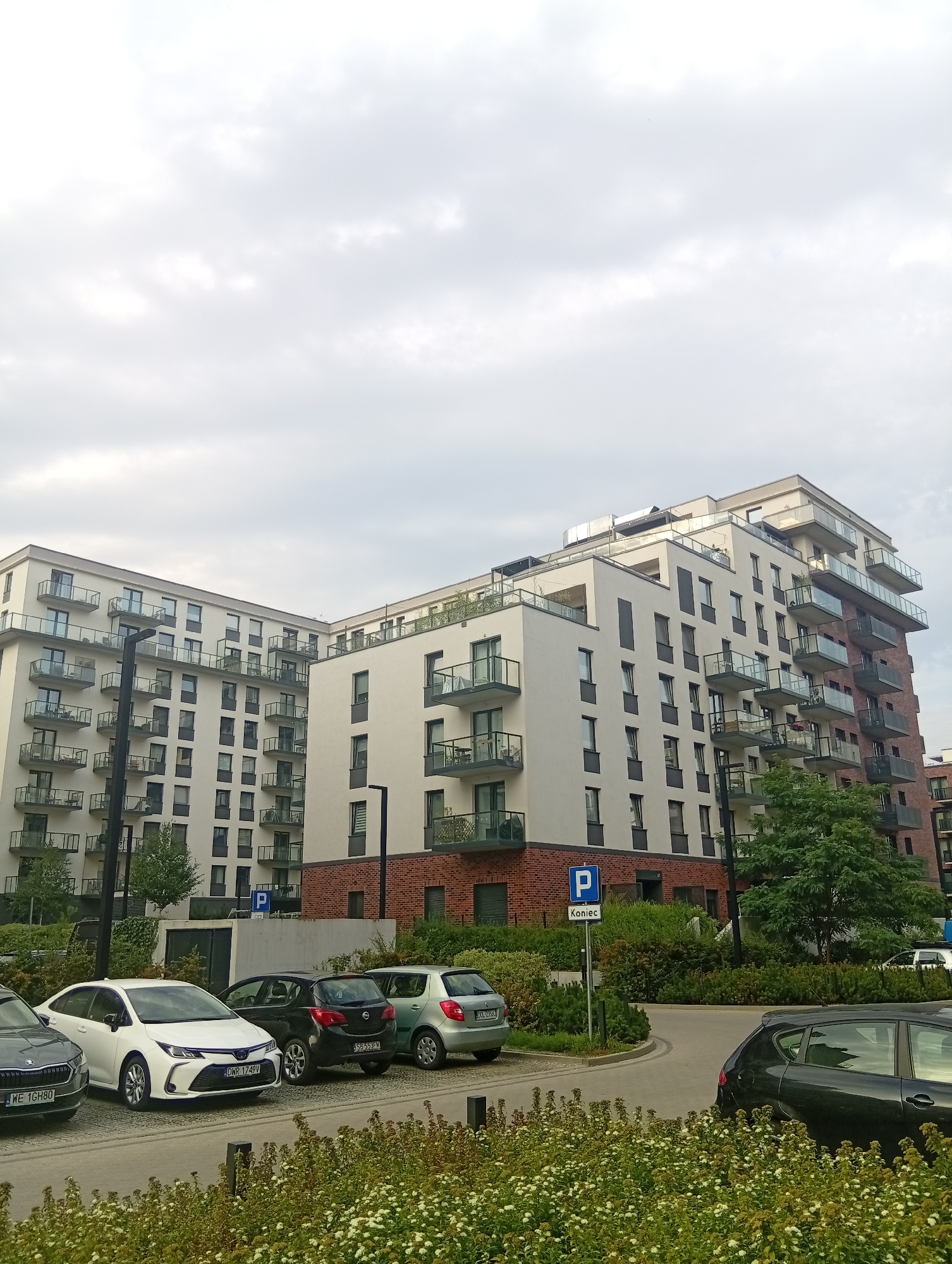 A medium shot captures a residential complex in Wrocław, Poland, featuring modern apartment buildings with white facades, numerous balconies with glass railings, and exposed brick accents. The buildings are multi-story, with some featuring a stepped architectural design. In the foreground, a paved parking area displays several cars, including a white sedan, a black hatchback, and a silver hatchback. A blue parking sign with the Polish word "Koniec" (End) is visible. Lush green trees and dense shrubbery surround the parking area and the buildings, with a bed of small white flowers in the immediate foreground. The sky above is overcast with diffused light.