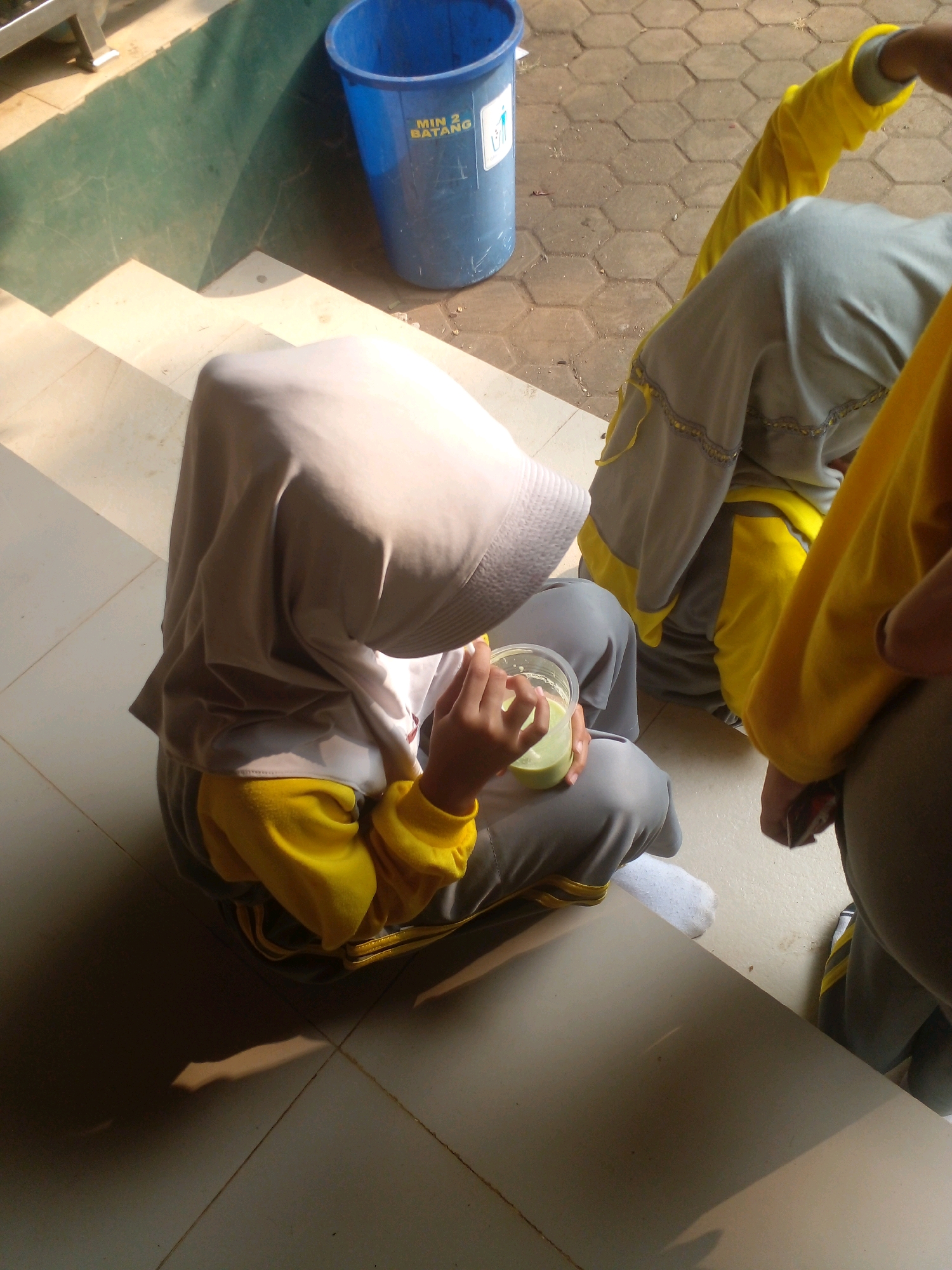 A person wearing a grey hijab and yellow long-sleeved shirt sits on a light-colored tiled step. They are holding a clear plastic cup containing a light green liquid and a straw. The person is also wearing grey pants and white socks. To the right, another person's legs are visible, also wearing grey pants with yellow trim. In the background, a large blue trash can with text "MIN 2 BATANG" is situated on a patterned pavement. Sunlight casts shadows on the steps and the pavement. The scene appears to be outdoors, likely on the grounds of a school in Banyuputih, Indonesia, given the school-related markings on the trash can.