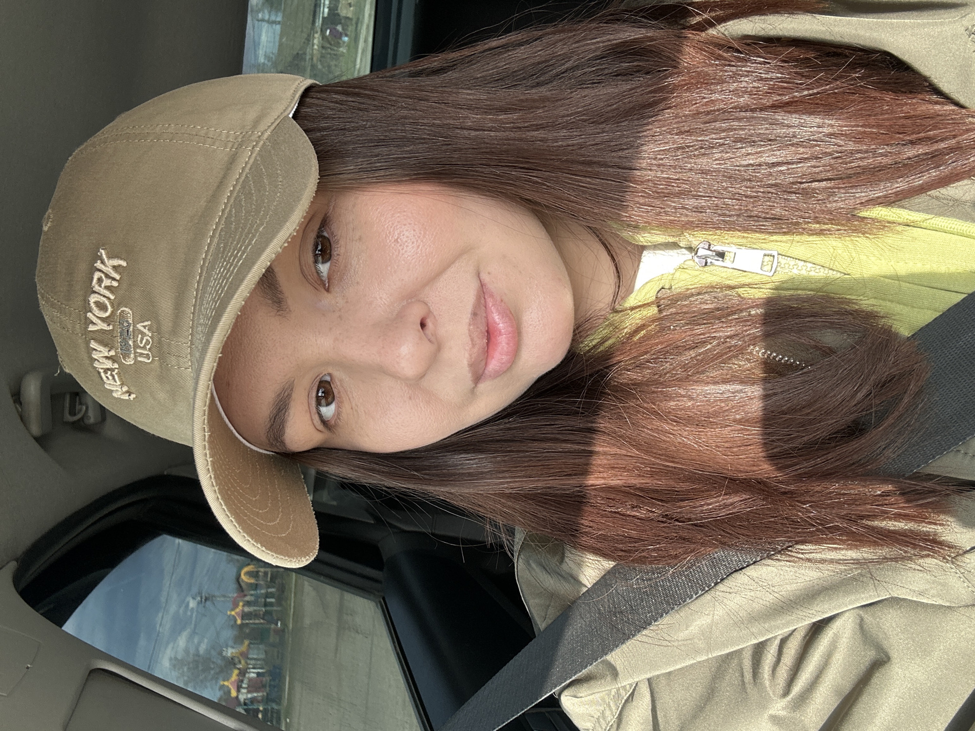 The media file displays a self-portrait or close-up photo of a young woman taken inside a car in Chaikovsky, Russia. The woman has long, straight dark brown hair, brown eyes, and light skin with visible freckles. She wears a light khaki-colored baseball cap and is dressed in a chartreuse green zipped top underneath an olive green jacket or coat. A dark grey seatbelt is visible across her chest.
She is looking directly at the camera with a subtle, gentle smile. Bright, direct sunlight illuminates her, creating strong highlights on her hair and casting distinct shadows across her forehead and parts of her hair and clothing. The interior of the car, including the headliner and window frame, is visible around her.
Through the car window, a clear blue sky and distant, somewhat blurry urban or recreational structures are discernible, suggesting an outdoor, daytime setting. The structures appear colorful, possibly indicative of amusement park rides or similar buildings in the Chaikovsky area.
The visible text "NEW YORK USA" is embroidered on the front of her baseball cap. The overall scene conveys a casual moment captured on a sunny day while traveling or parked.